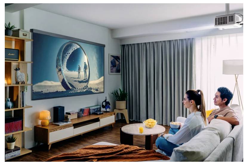 A man and woman sit on a sofa watching a projection screen showing an image of two people walking on a sandy surface with a circular frame.