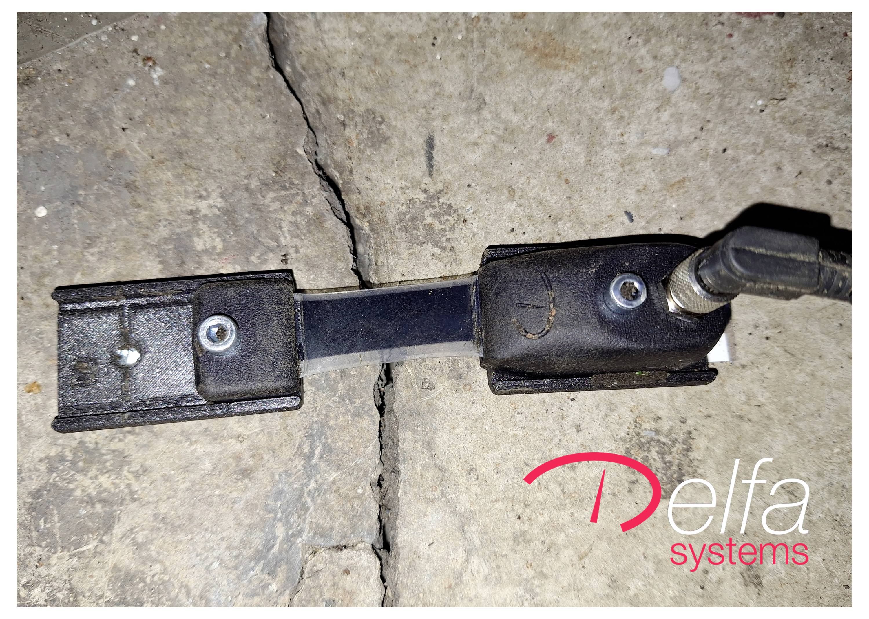 An electronic sensor mounted on a crack in a concrete surface, with the 'Delfa systems' logo in the lower right corner.