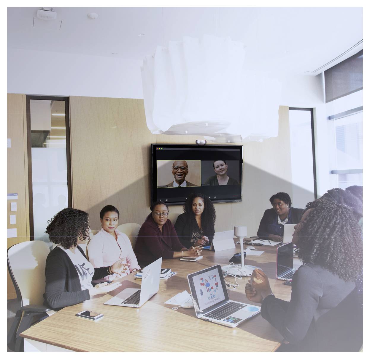 A group of people in a conference room, engaged in a meeting. A video call is displayed on the screen with two participants on it.