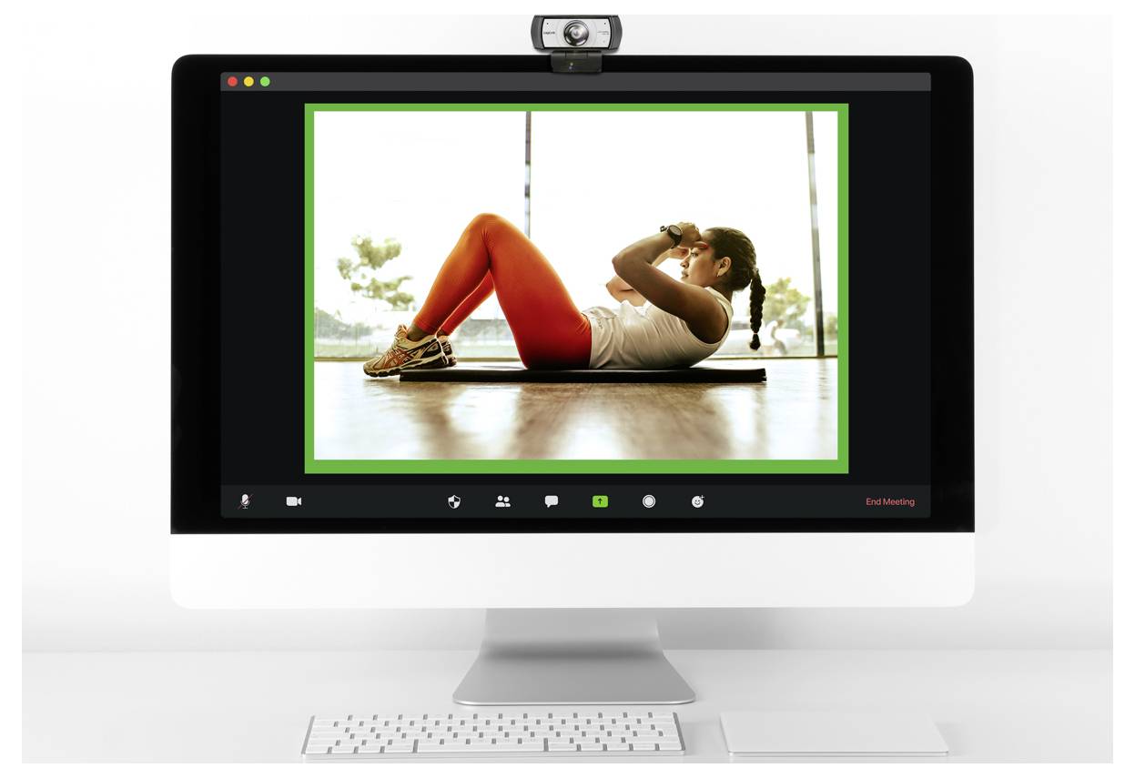 A computer screen shows a video call with a person doing sit-ups on a mat, wearing red pants and a white top, indoors with a bright background.