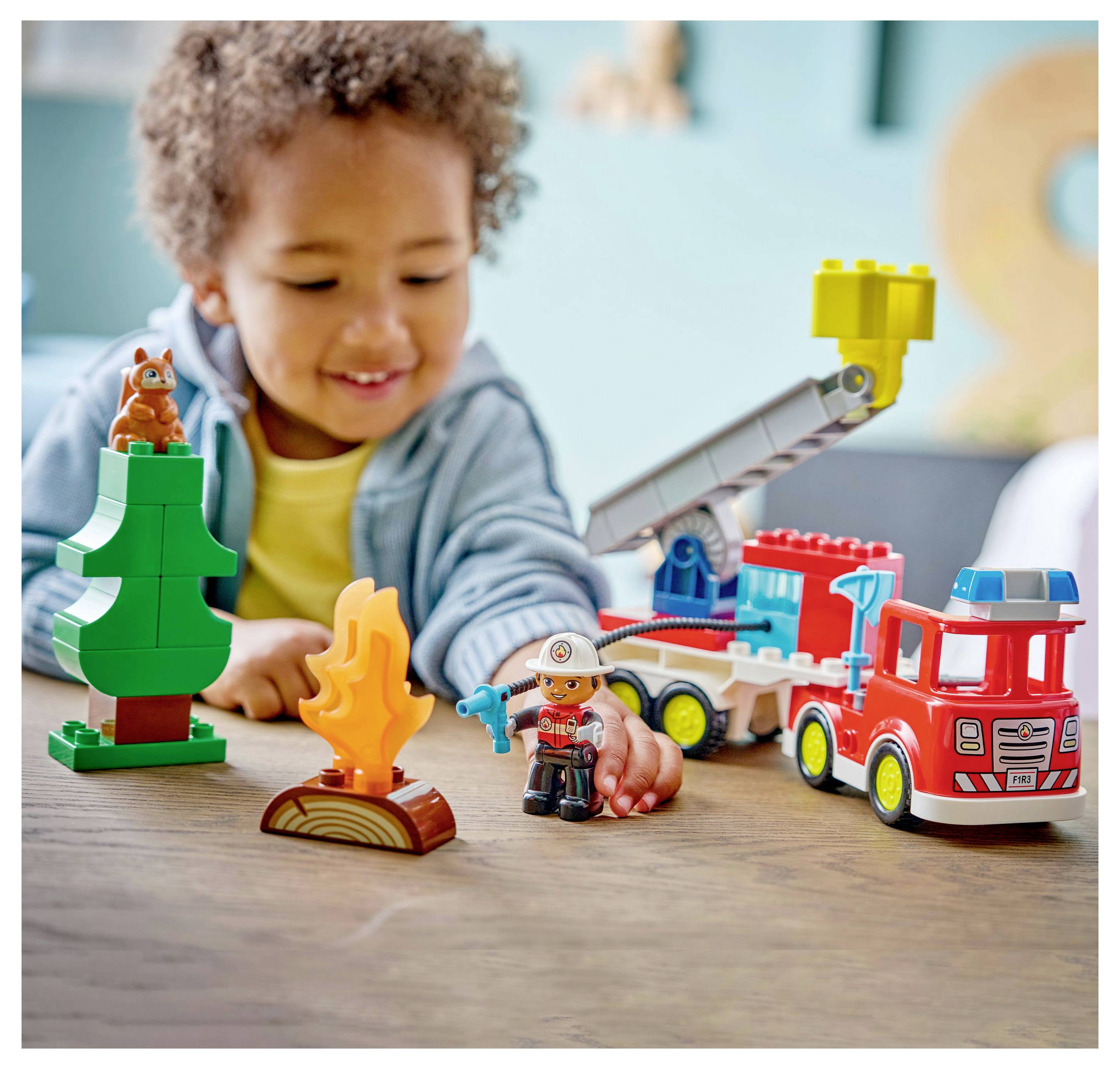 A child plays with building blocks and toys, including a fire truck, tree, and small figurines, in a colorful, indoor setting.