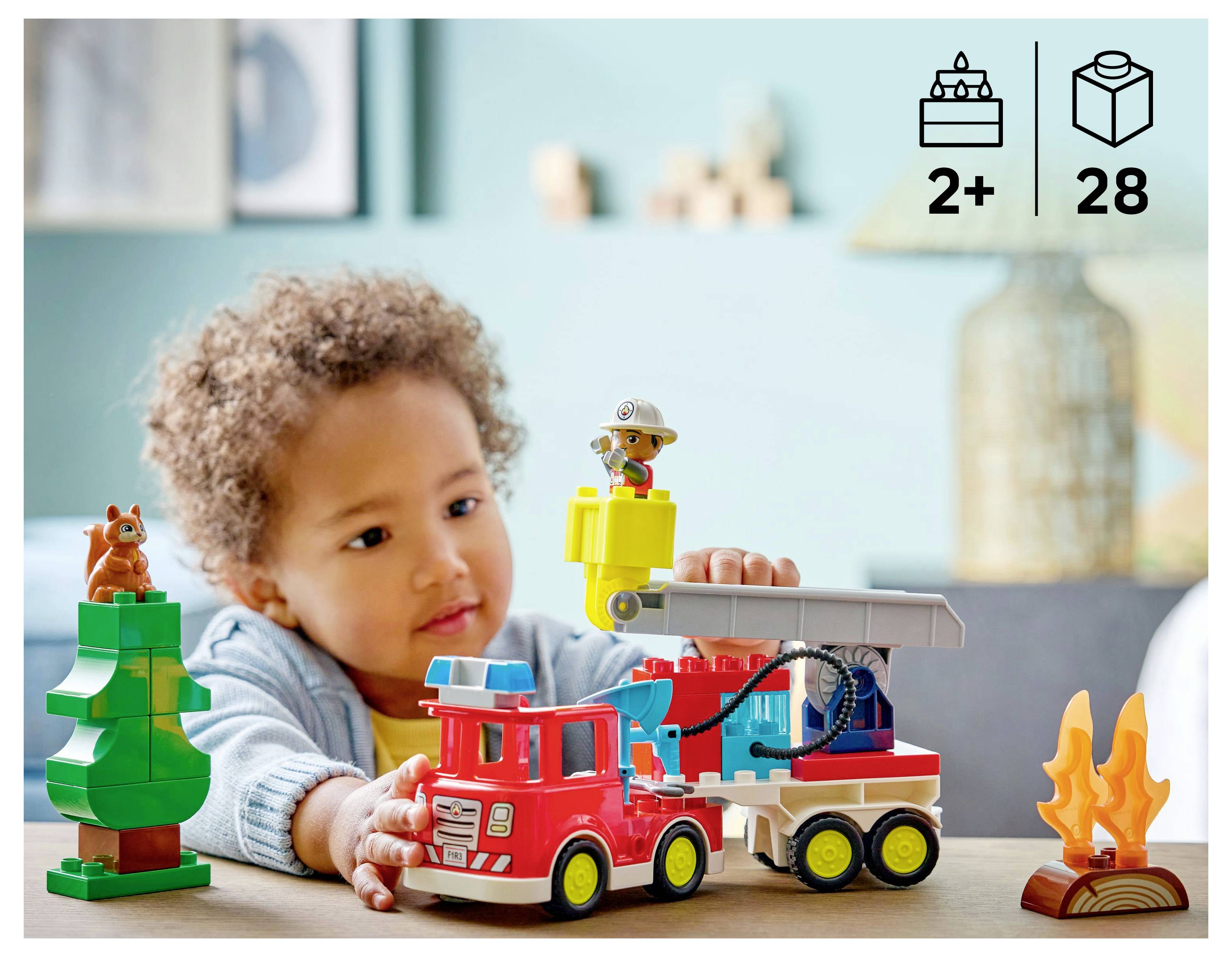 A child plays with a toy fire truck set, including a tree, fireman figure, and flames, suitable for ages 2 and up with 28 pieces.