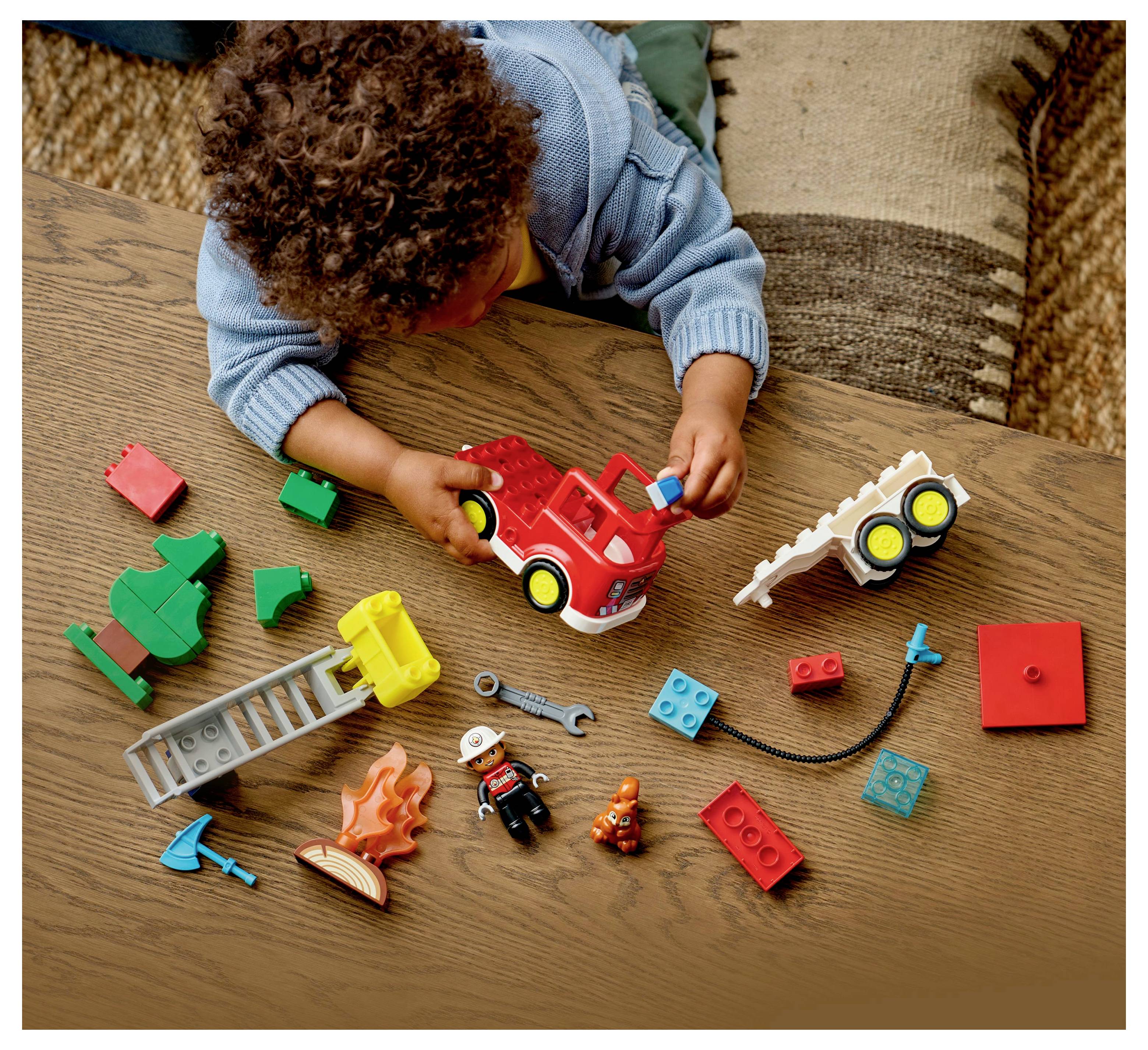 A child plays on a wooden surface with a red toy fire truck and various colorful toy blocks and figures scattered around.