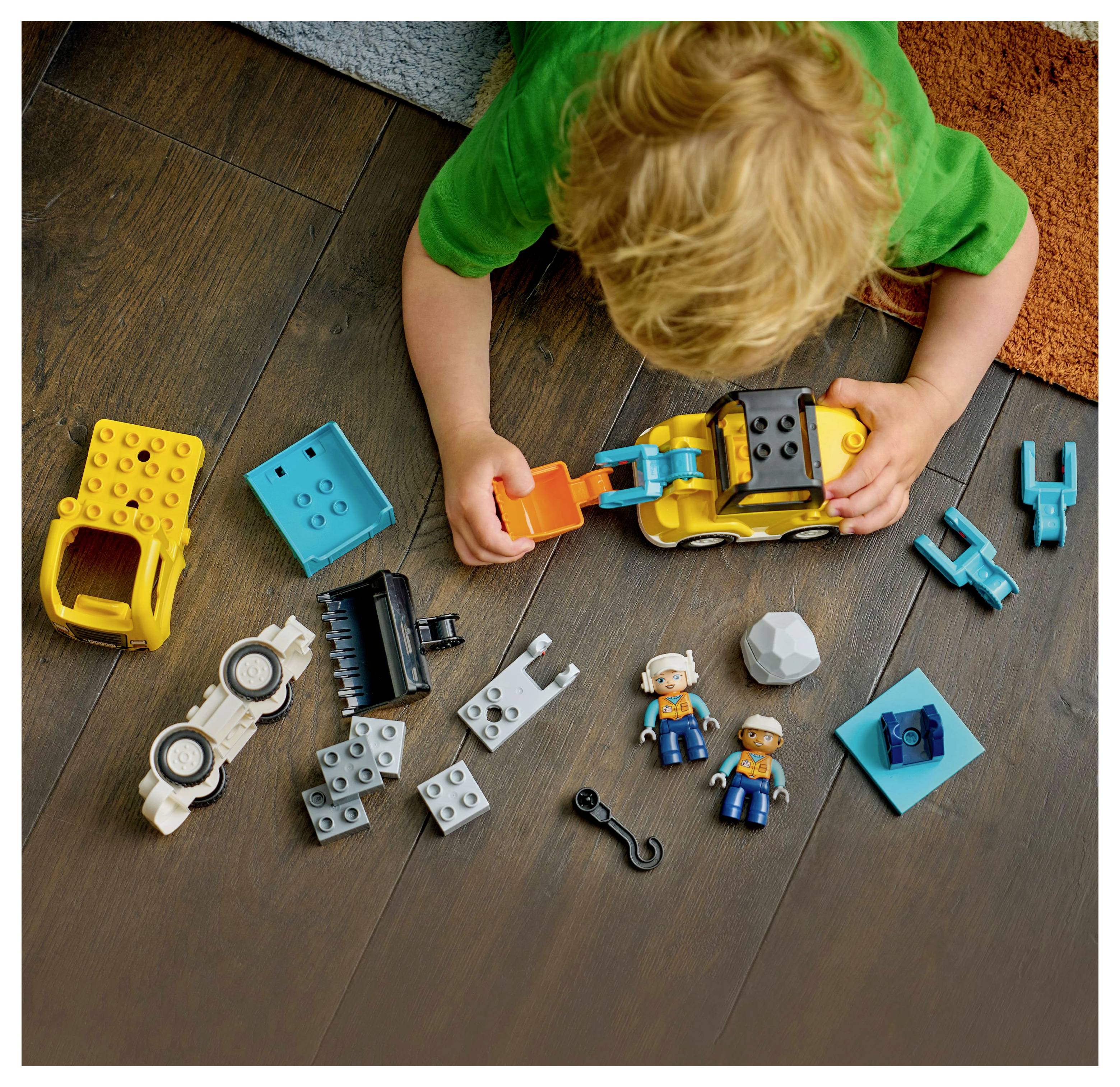 A child in a green shirt plays with colorful toy building blocks and figures on a wooden floor.