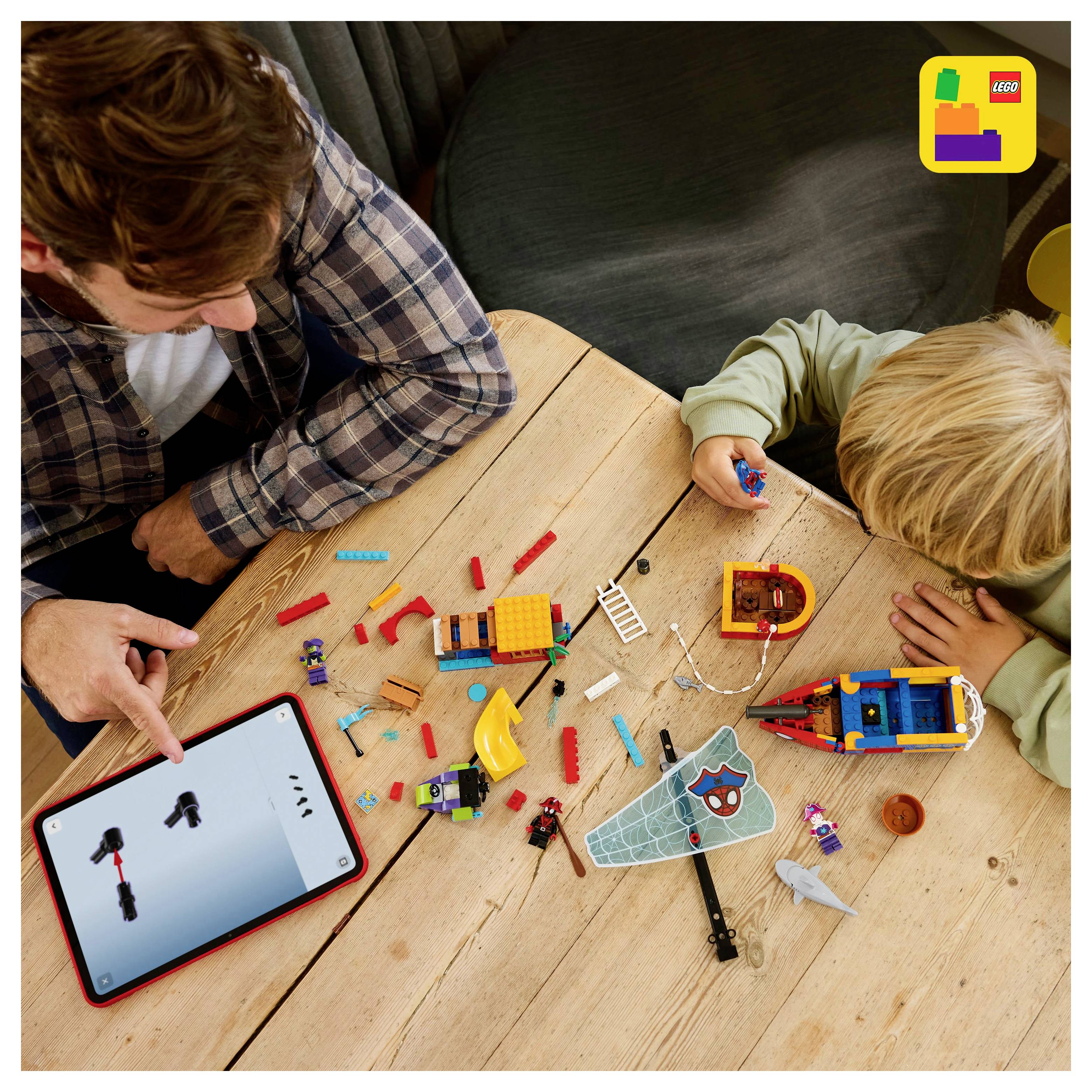 A child and adult use a tablet with building instructions to assemble LEGO bricks on a wooden table, creating a toy spaceship and mini-figures.