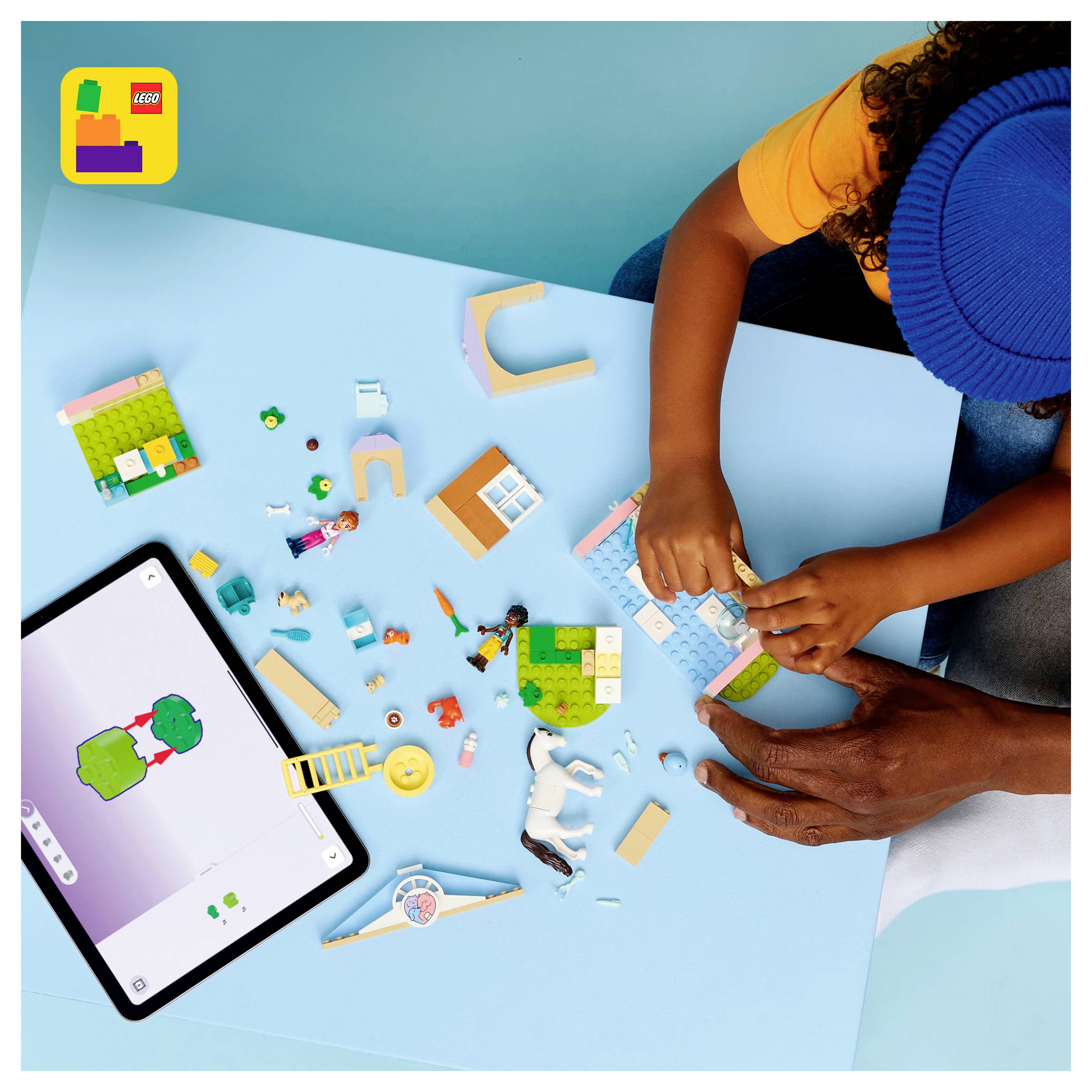 A child and adult assemble LEGO pieces on a blue surface beside a tablet displaying a LEGO app. The setting suggests interactive play.