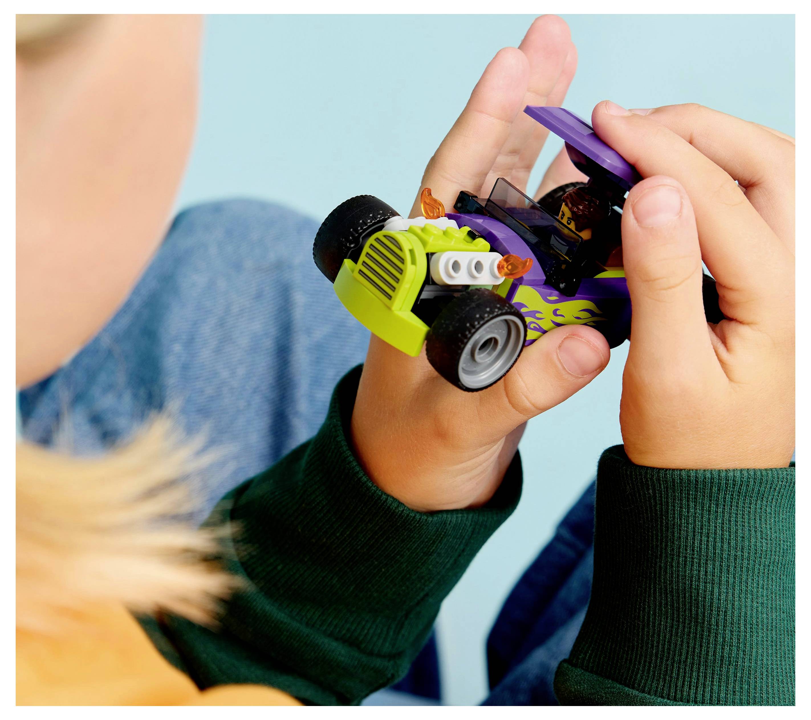 A child holds a small toy car with a vibrant green grill and flame decals. The child wears a green sweater and the background is light blue.
