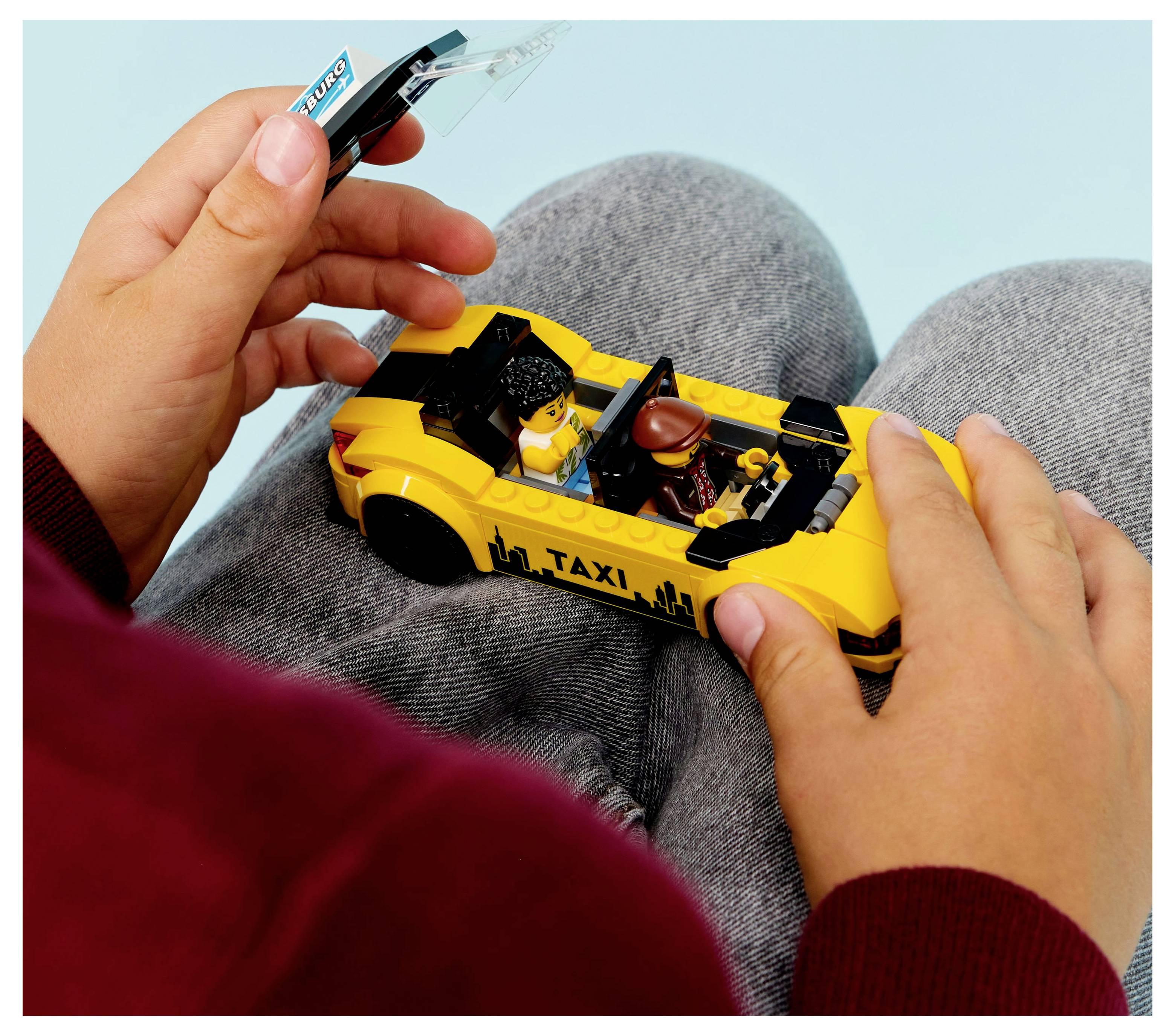 A child holds a LEGO taxi car model in their hands. The taxi has mini figures inside and the setting appears casual and playful.