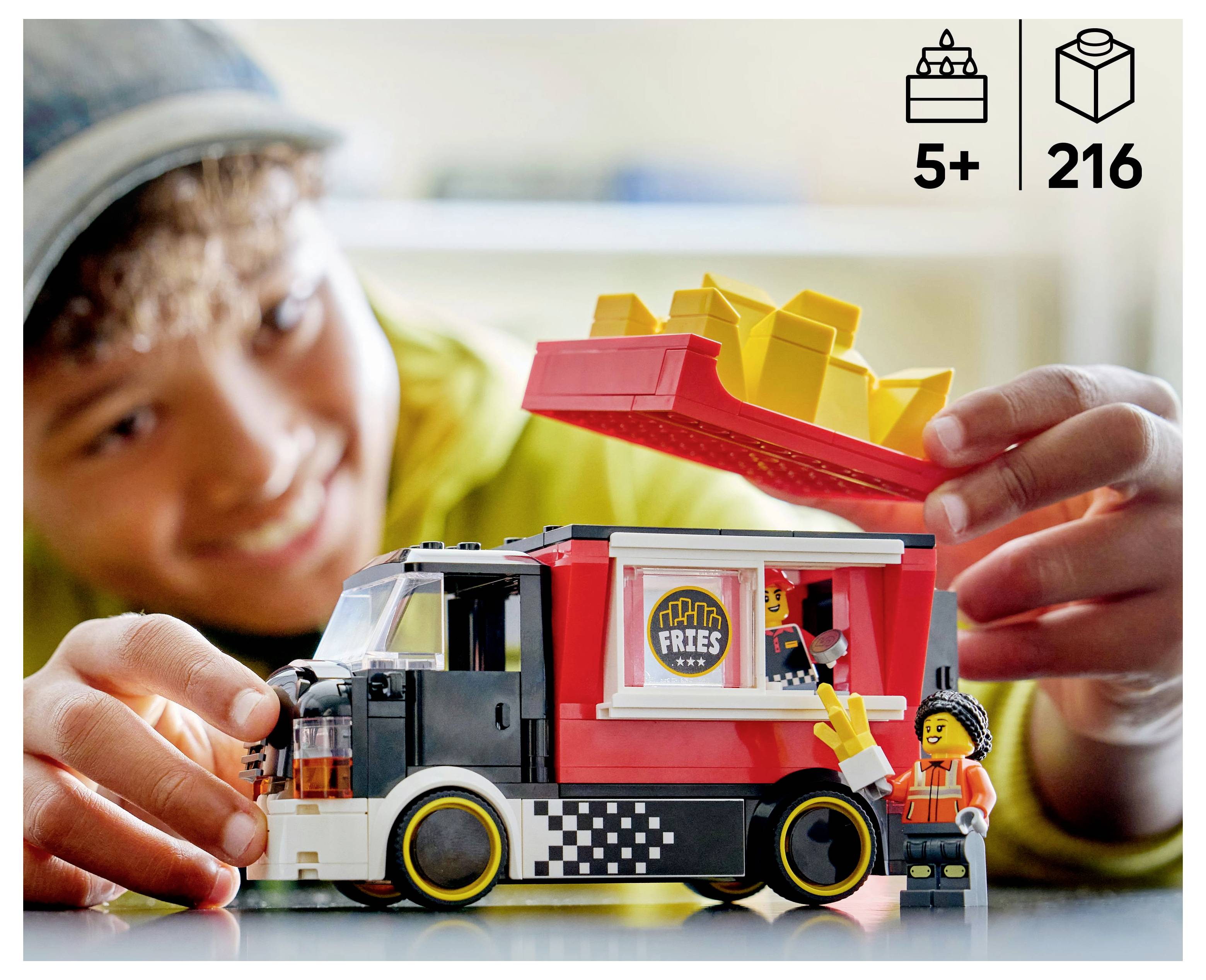 A child plays with a toy food truck set, featuring a 'Tasty Fries' sign, removable roof with bricks resembling fries, and a minifigure.