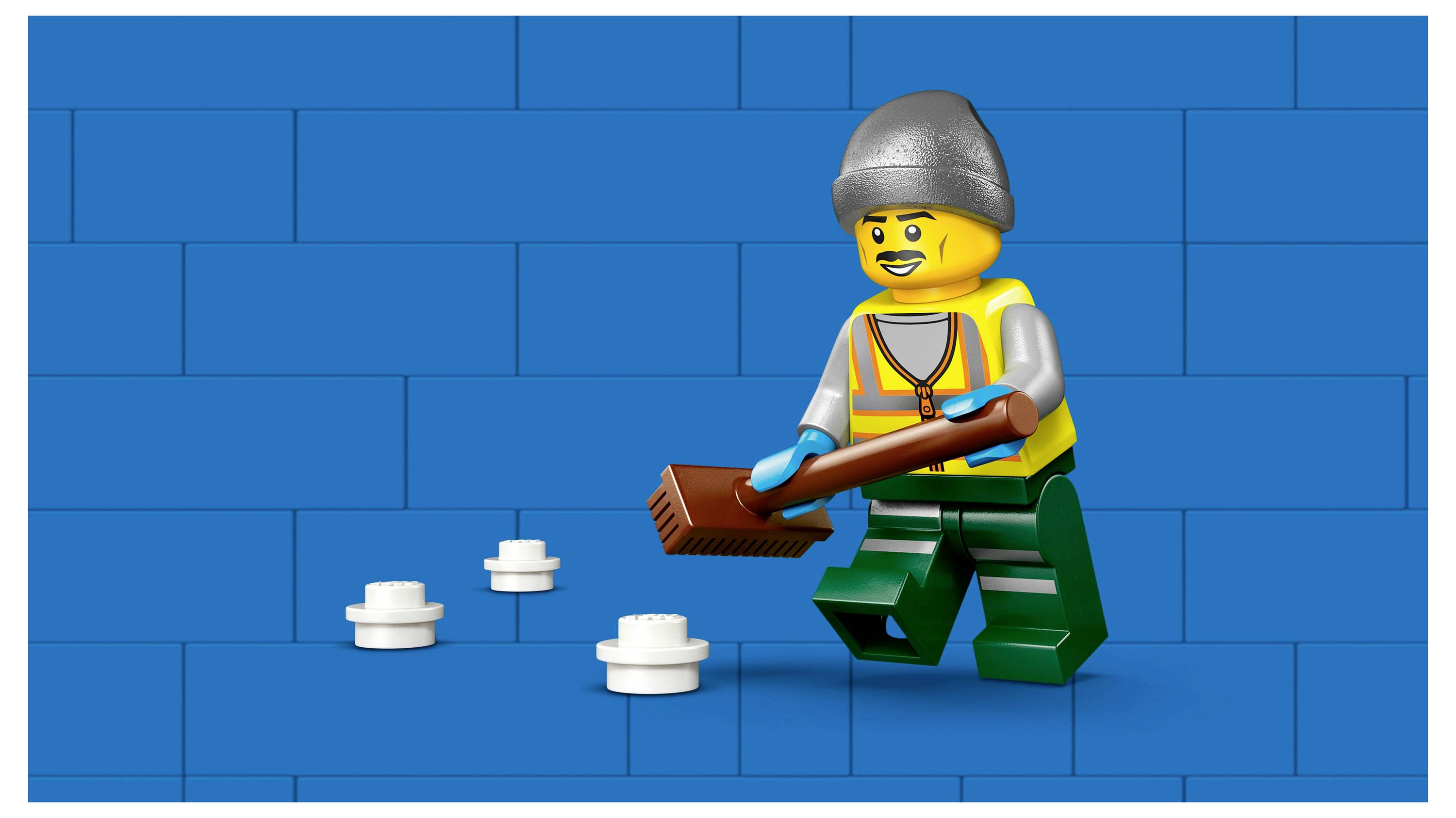 A LEGO construction worker in a hat and safety vest is sweeping a blue brick floor. Several white LEGO bricks are scattered nearby.