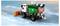 A toy snow plow truck on a snowy road dispenses snowballs. The truck is green and white with an orange plow on the front.