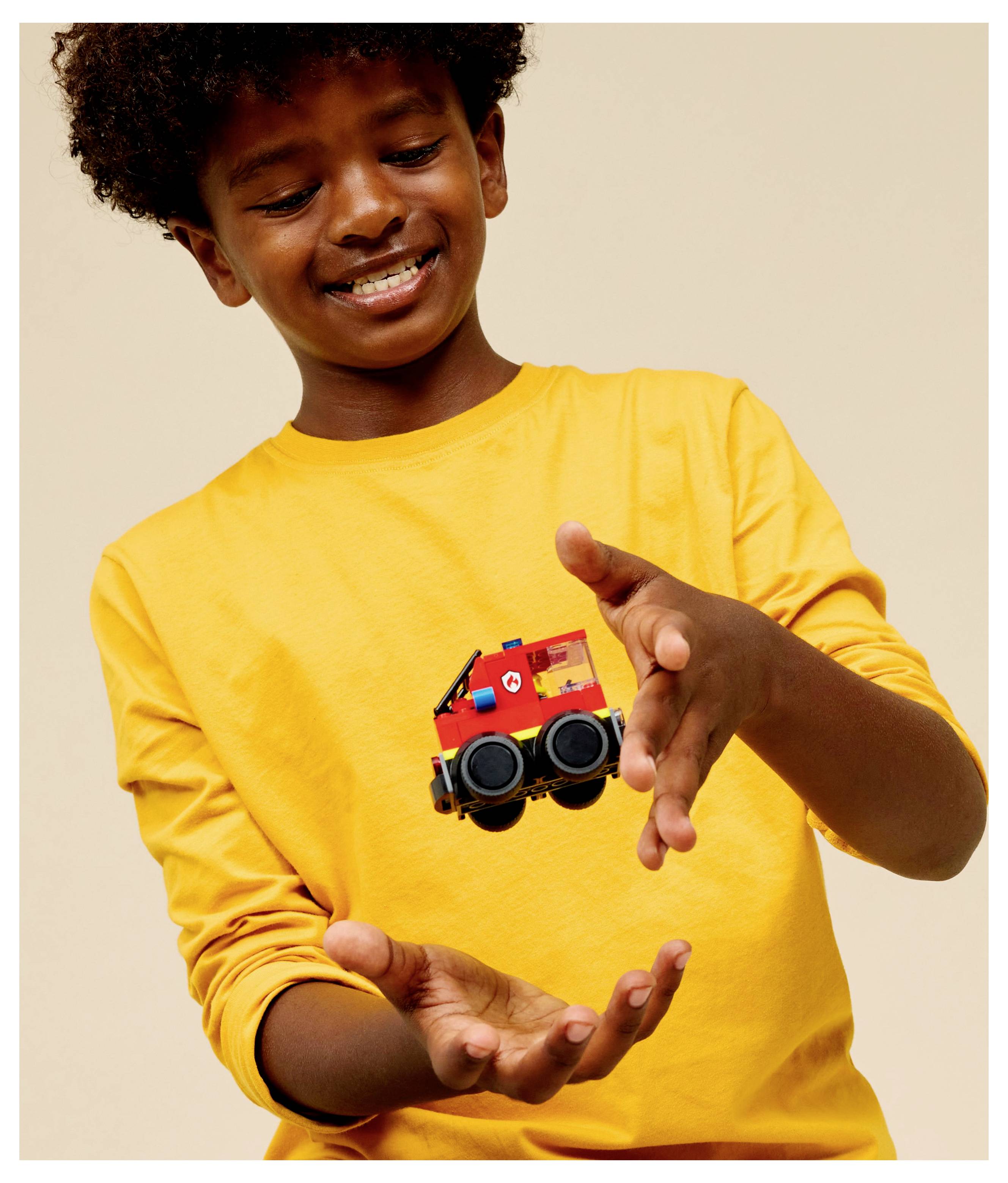 A child joyfully wearing a yellow shirt with a small red toy truck design on it, playfully engaging with it against a neutral background.