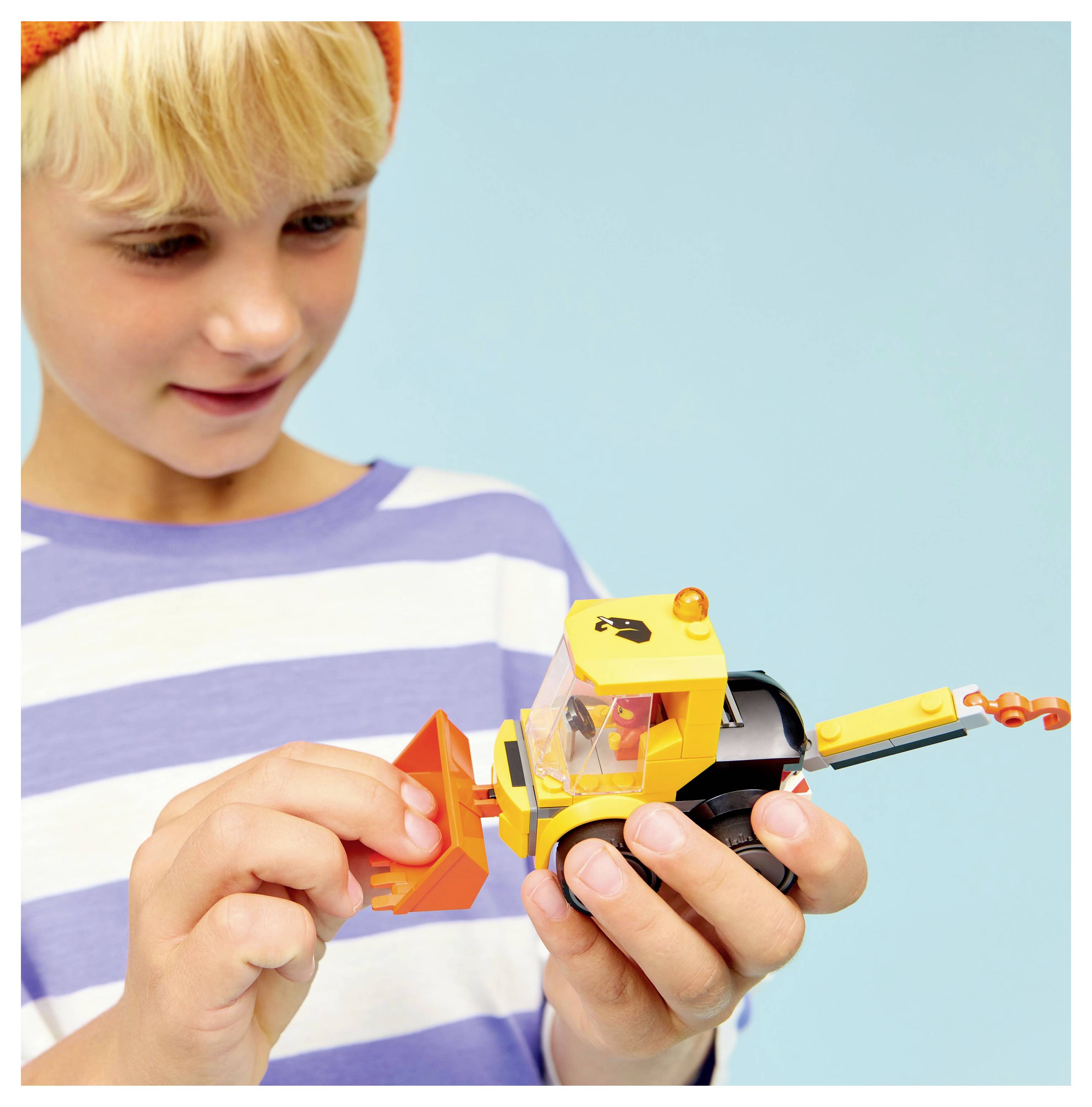 A child in a striped shirt and orange hat plays with a yellow toy truck. The child focuses on attaching a small part to the truck.