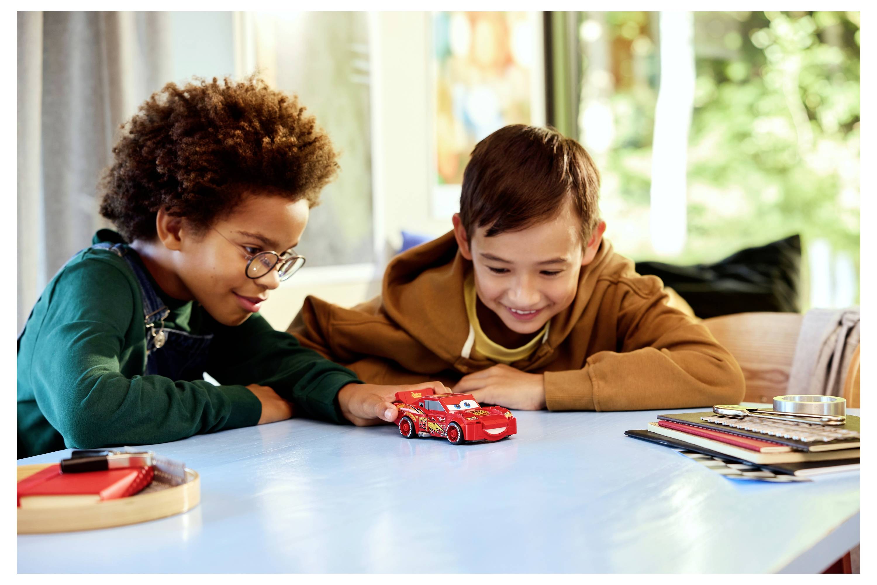 Two children happily playing with a red toy car on a table, surrounded by art supplies, in a bright room with green trees visible outside.