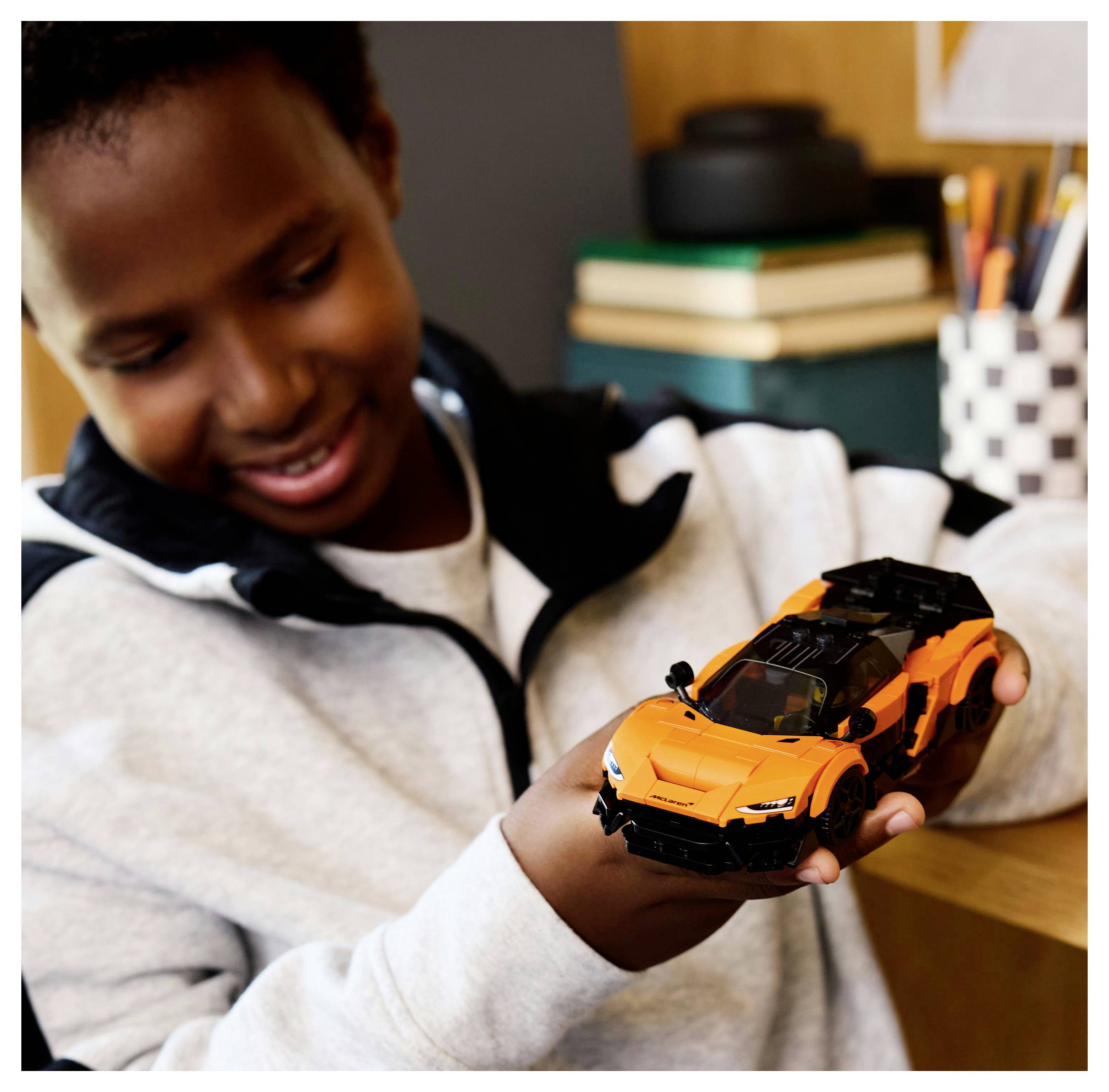 A person smiling while holding a detailed toy car model inside a room. Books and pencils are visible on a desk in the background.