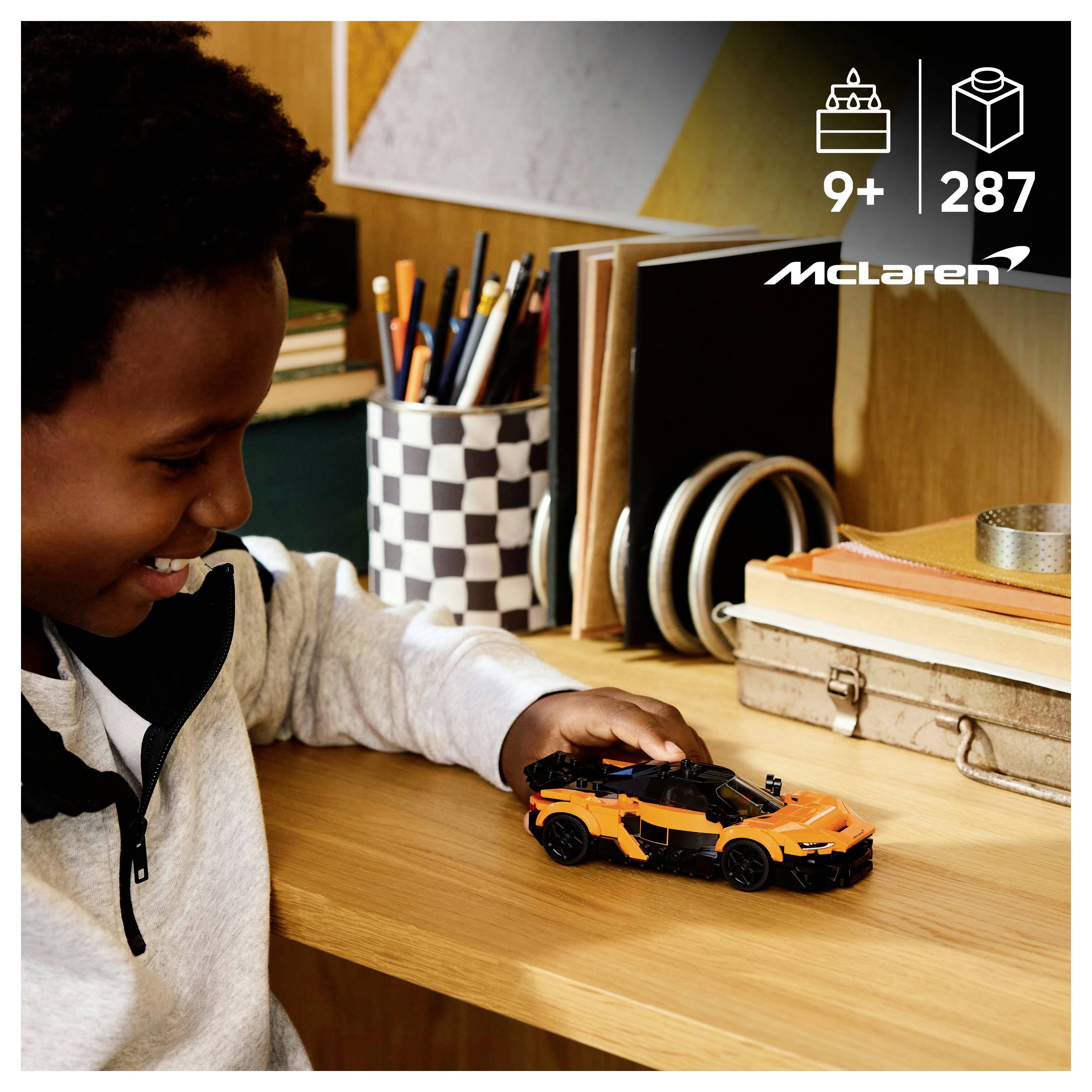 A child plays with an orange toy car on a wooden desk. Books and a pencil holder are in the background.