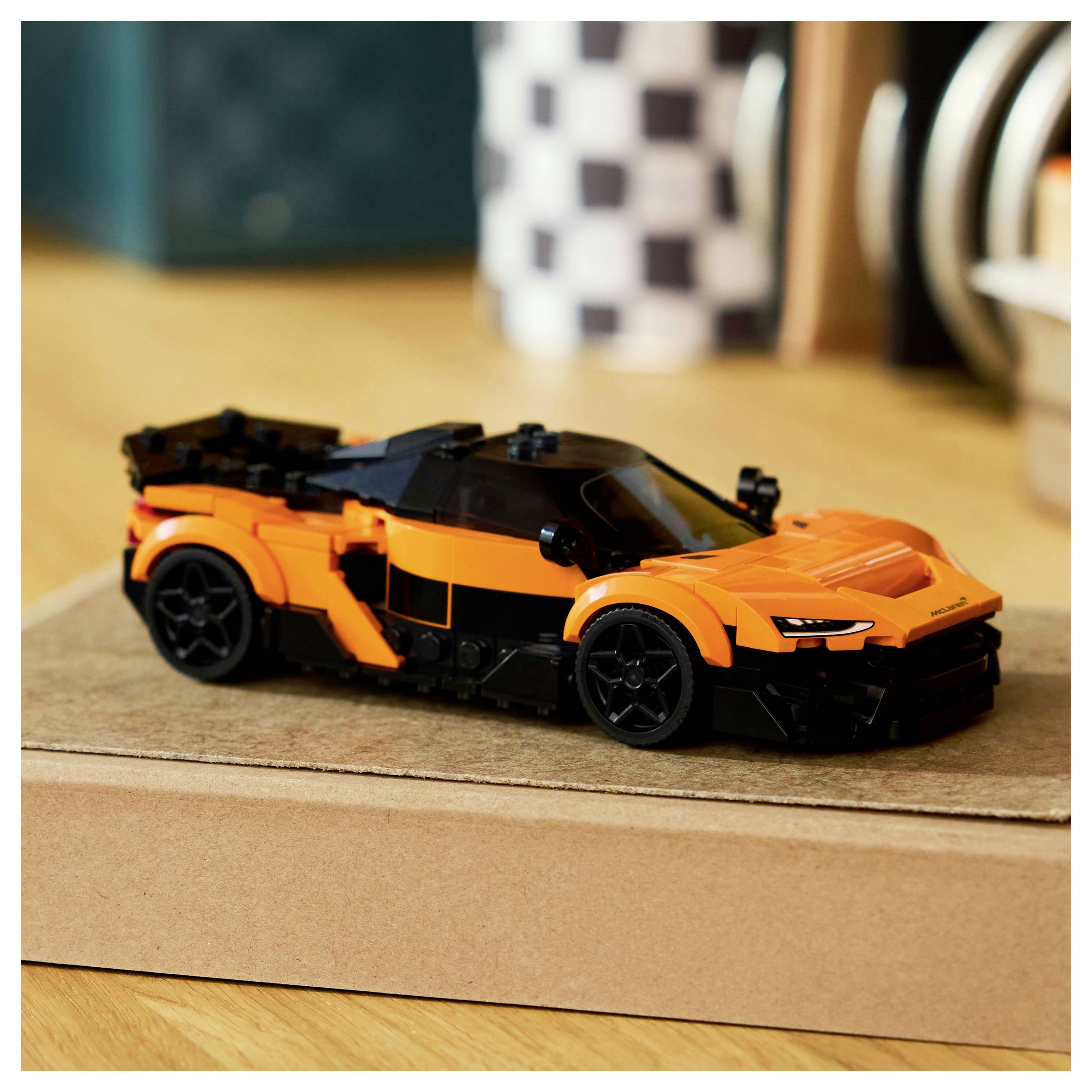 A small orange toy car with black details is placed on a cardboard box, set on a wooden floor against a blurred background.