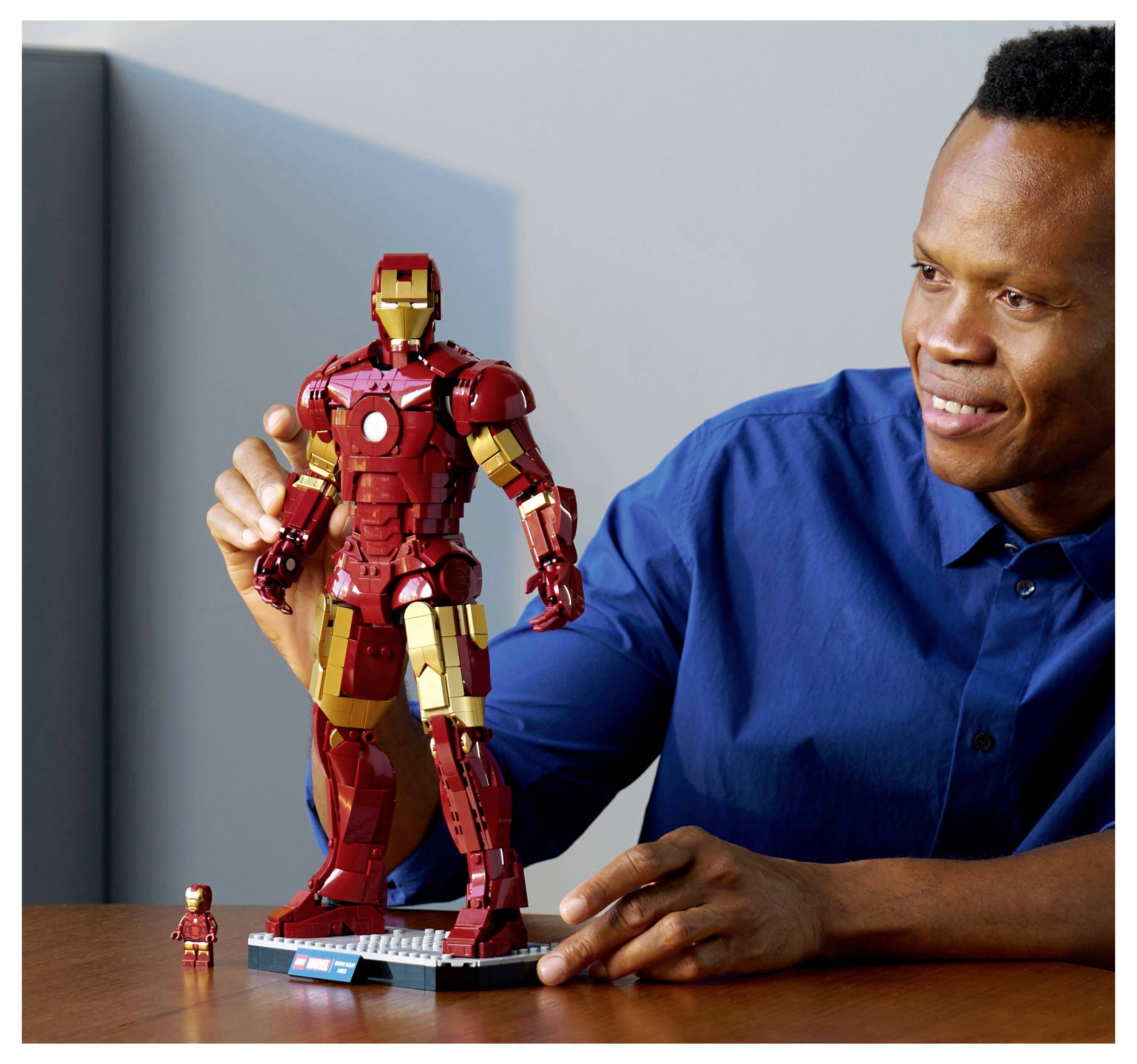 A person in a blue shirt smiles while holding a large red and gold action figure on a table, with a smaller matching figure beside it.