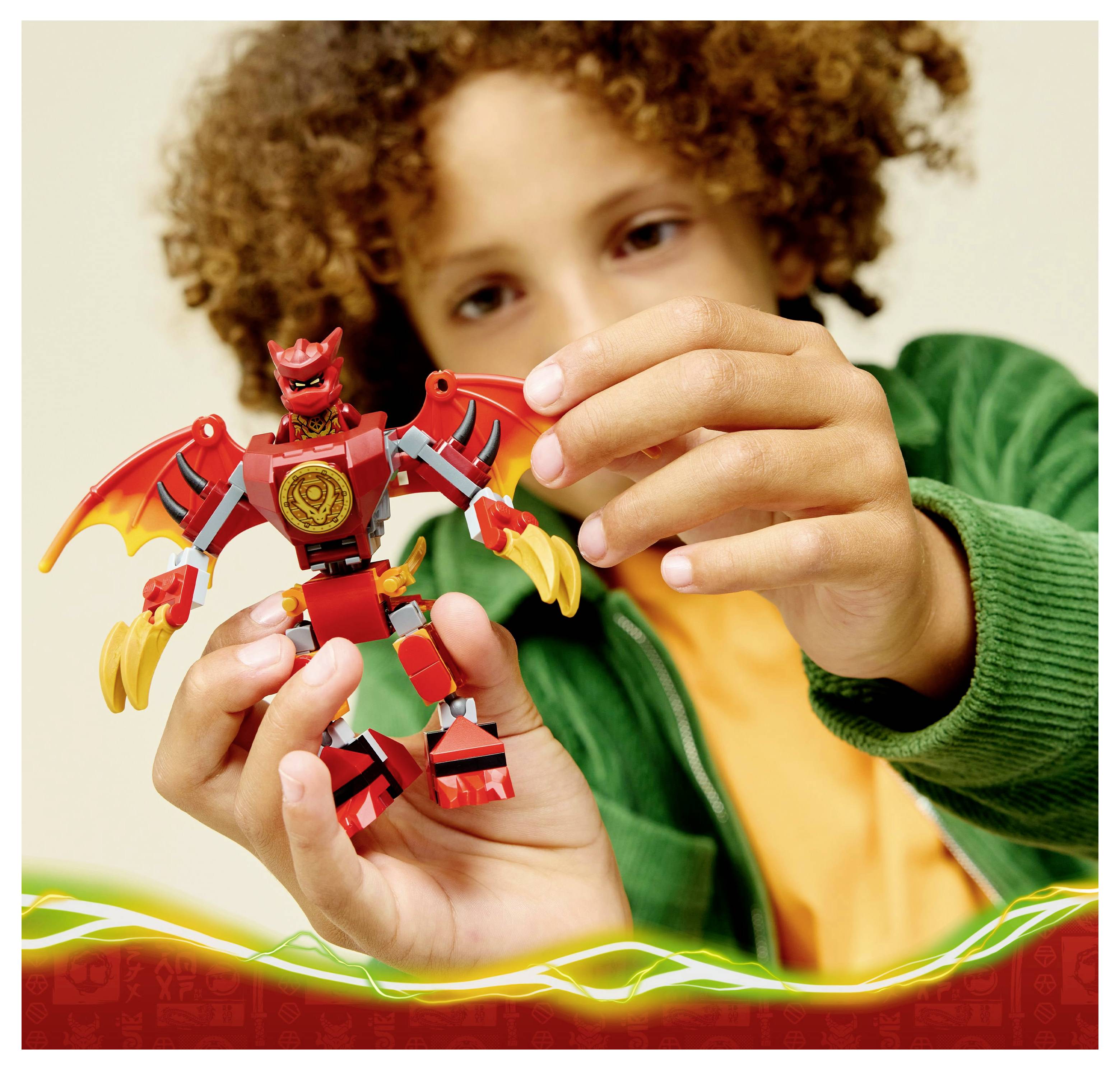A child in a green jacket holds and examines a small red and yellow dragon-themed action figure with wings and claw-like hands.