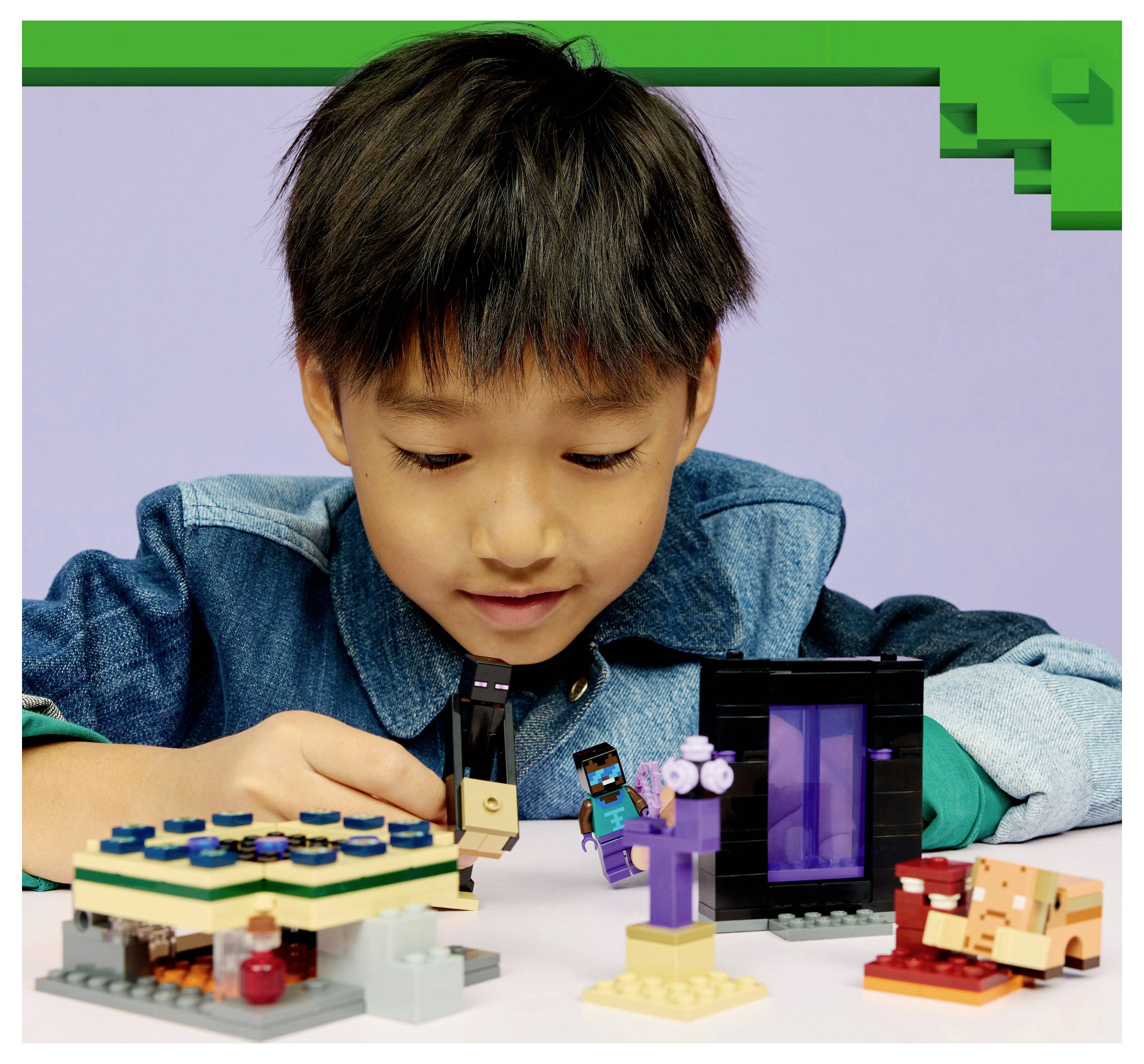 A child in a blue shirt is playing with Minecraft-themed LEGO, building small structures and figures on a table against a purple backdrop.