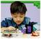 A child in a blue shirt is playing with Minecraft-themed LEGO, building small structures and figures on a table against a purple backdrop.