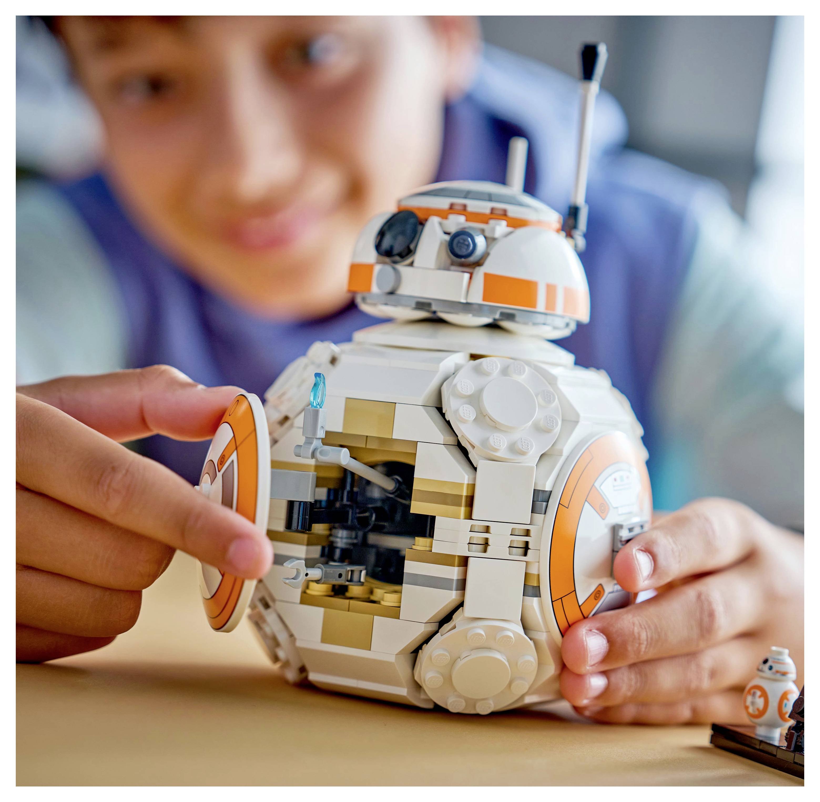 A child plays with a toy robot on a table, focusing intently on its details. The robot is white with orange accents, resembling a droid.