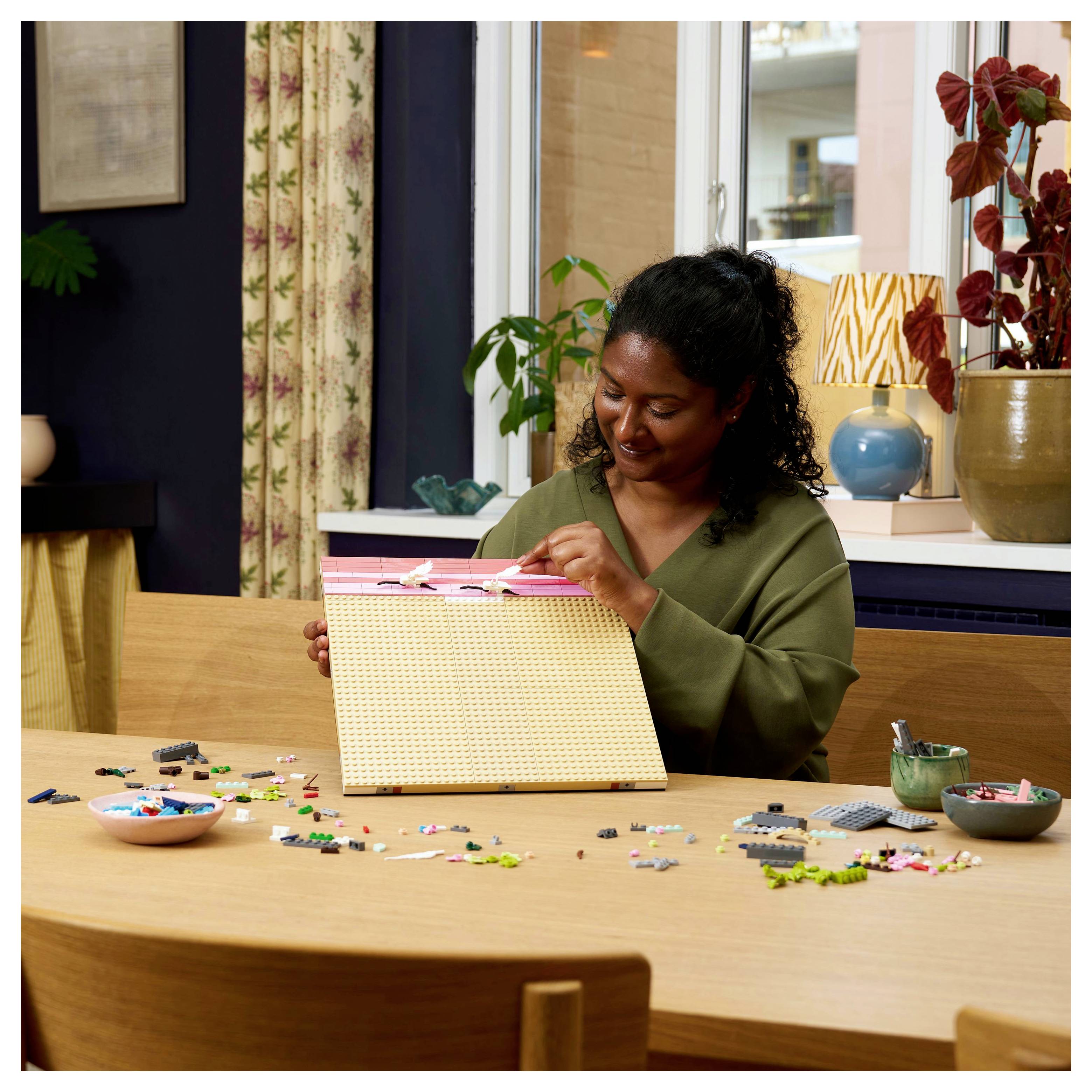 A person sits at a table assembling a colorful block art piece, surrounded by scattered blocks and a plant in the background.
