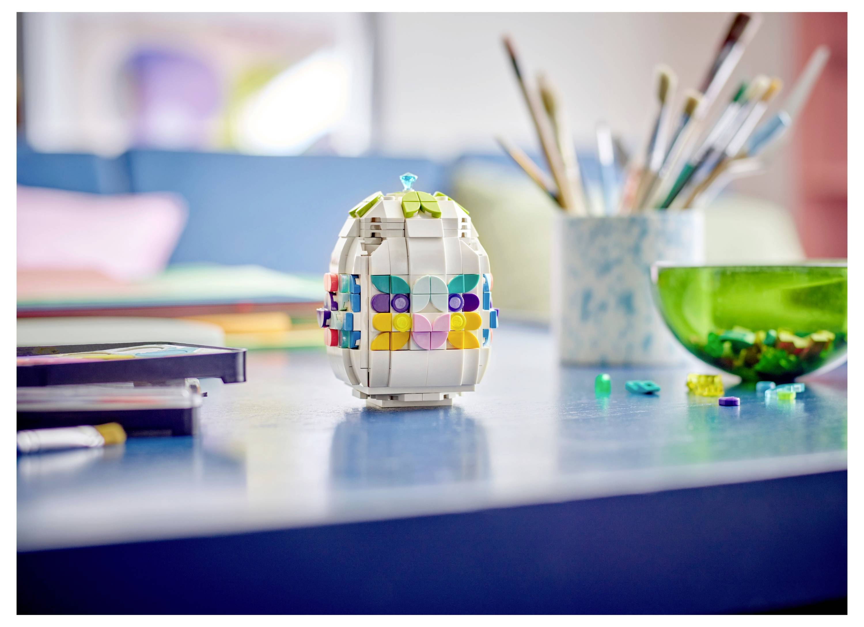 A colorful block toy sits on a table surrounded by paintbrushes, a green bowl, and art supplies, suggesting a creative setting.
