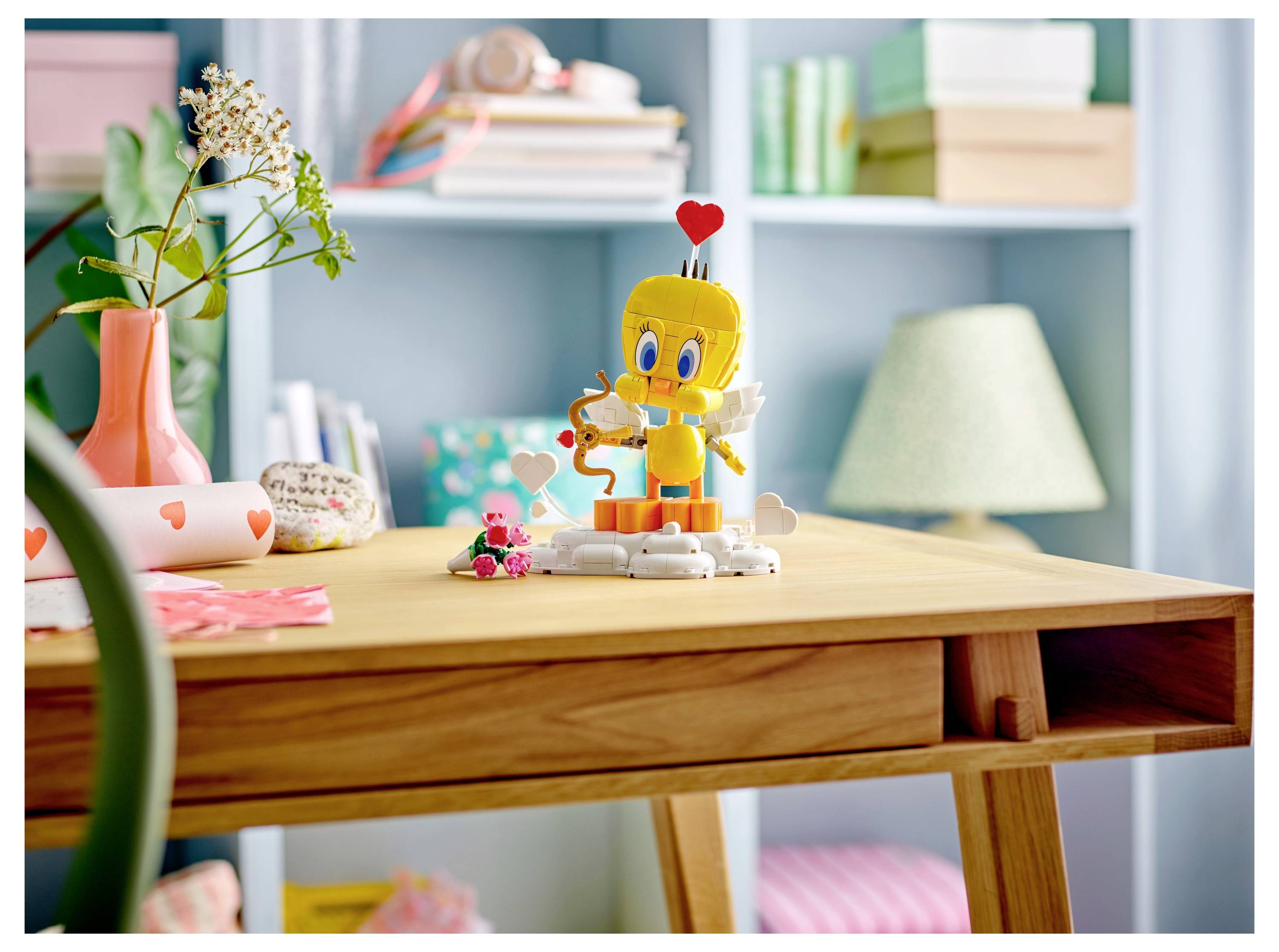 A workspace with a wooden desk, decorated with a small bird toy holding a heart, and a heart-themed vase with flowers, set against a bookshelf.