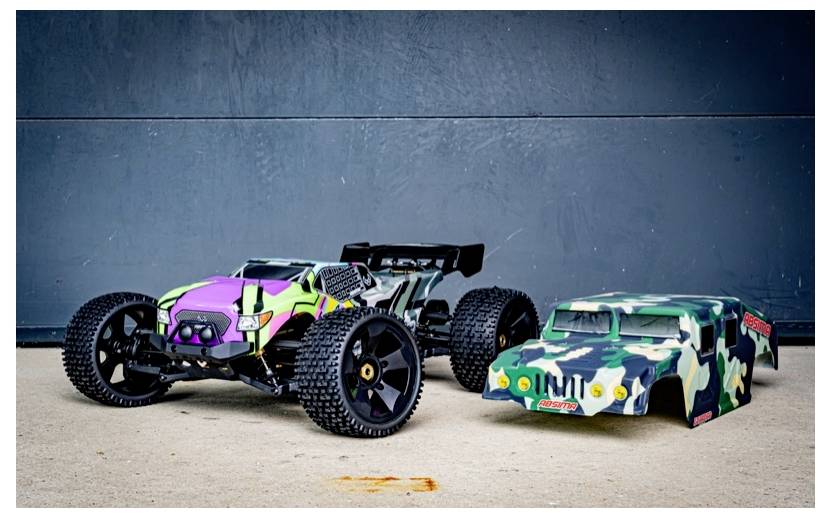A remote-controlled car with black wheels and a camouflage body stands on concrete, next to a similar detached car shell.