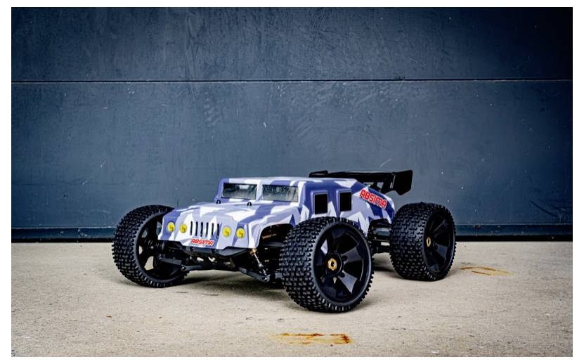 A remote-controlled, camouflage-patterned car with large black tires is positioned against a dark, textured wall.
