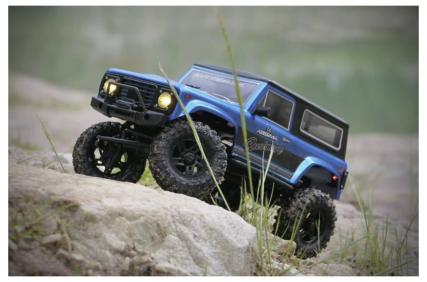 A blue toy off-road vehicle with large tires drives over a rocky terrain, showcasing its suspension and rugged design.