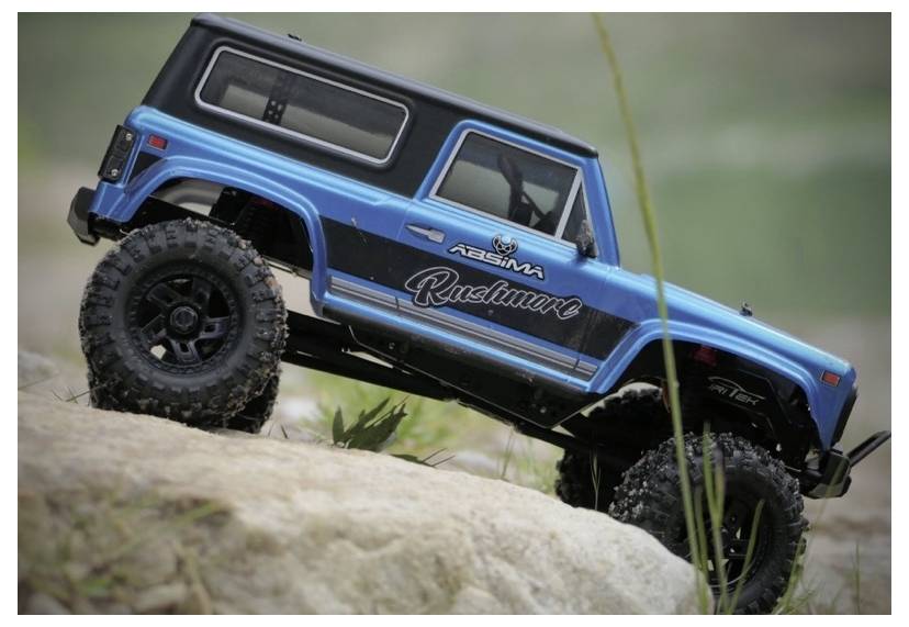 A blue remote-controlled truck, branded with 'Rushmore', climbs over a rocky terrain, demonstrating its off-road capabilities.