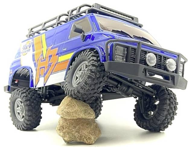 A blue toy off-road vehicle with large tires and decals is climbing over stacked rocks, demonstrating its suspension and traction.