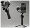 Two gimbals displayed: one holds a camera with flash on, the other holds a smartphone. Both are mounted on tripods in a well-lit studio.