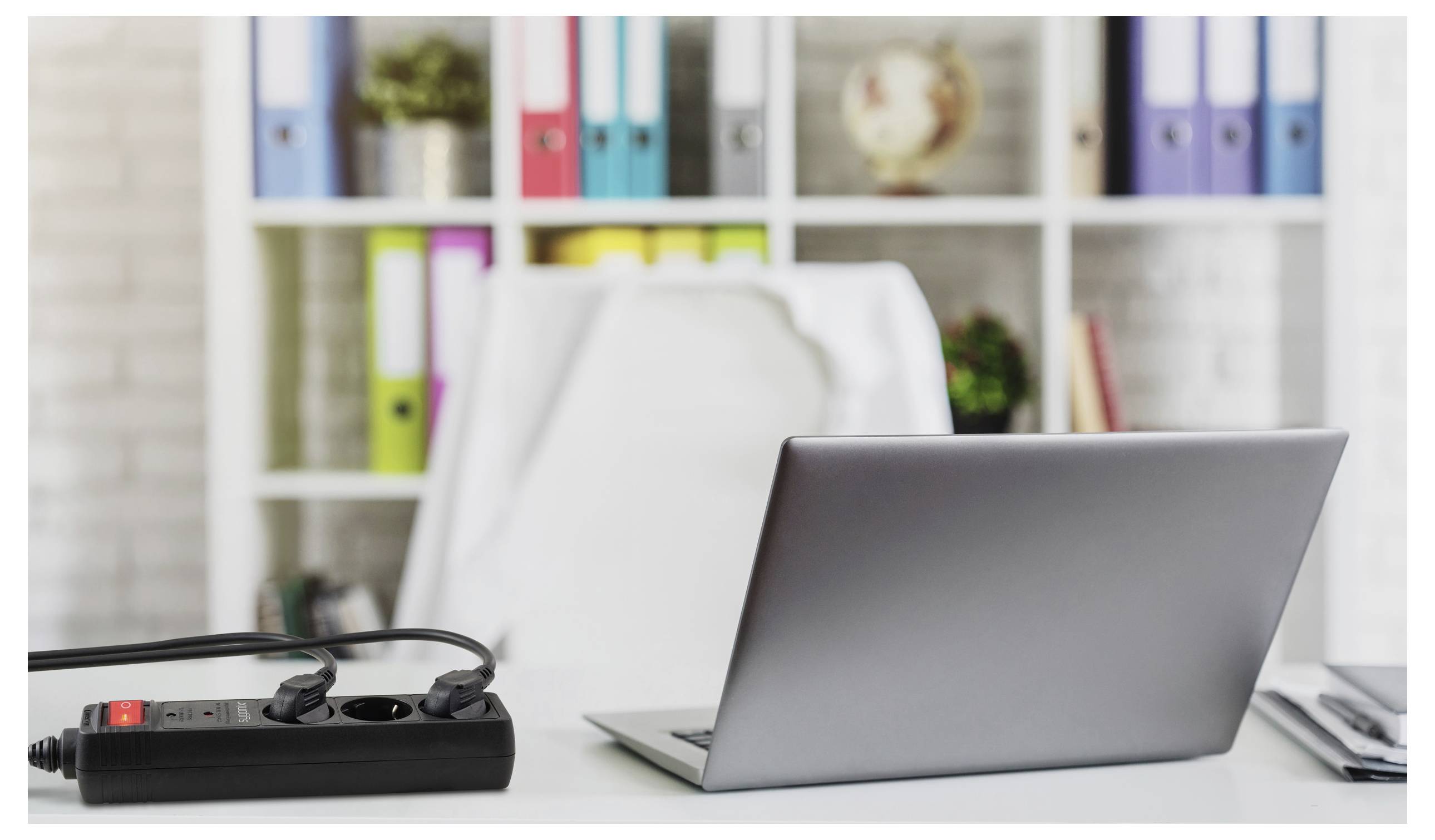 'Laptop on a desk with a power strip, in front of shelves with colorful binders and a globe, suggesting a home office setup.'