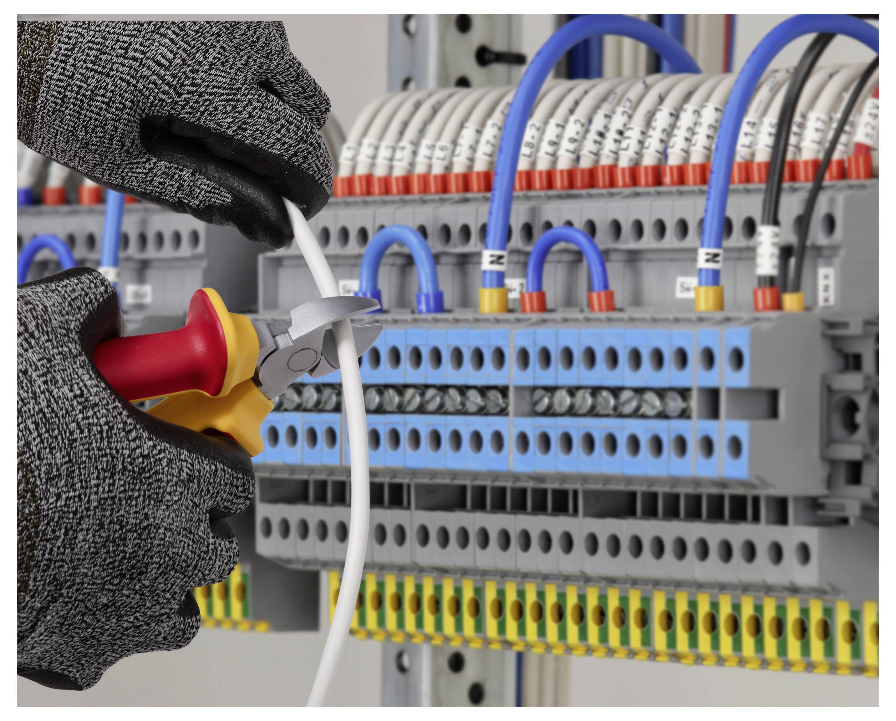 Gloved hands use wire cutters to strip a cable inside an electrical control panel with terminal blocks and wiring.<br>