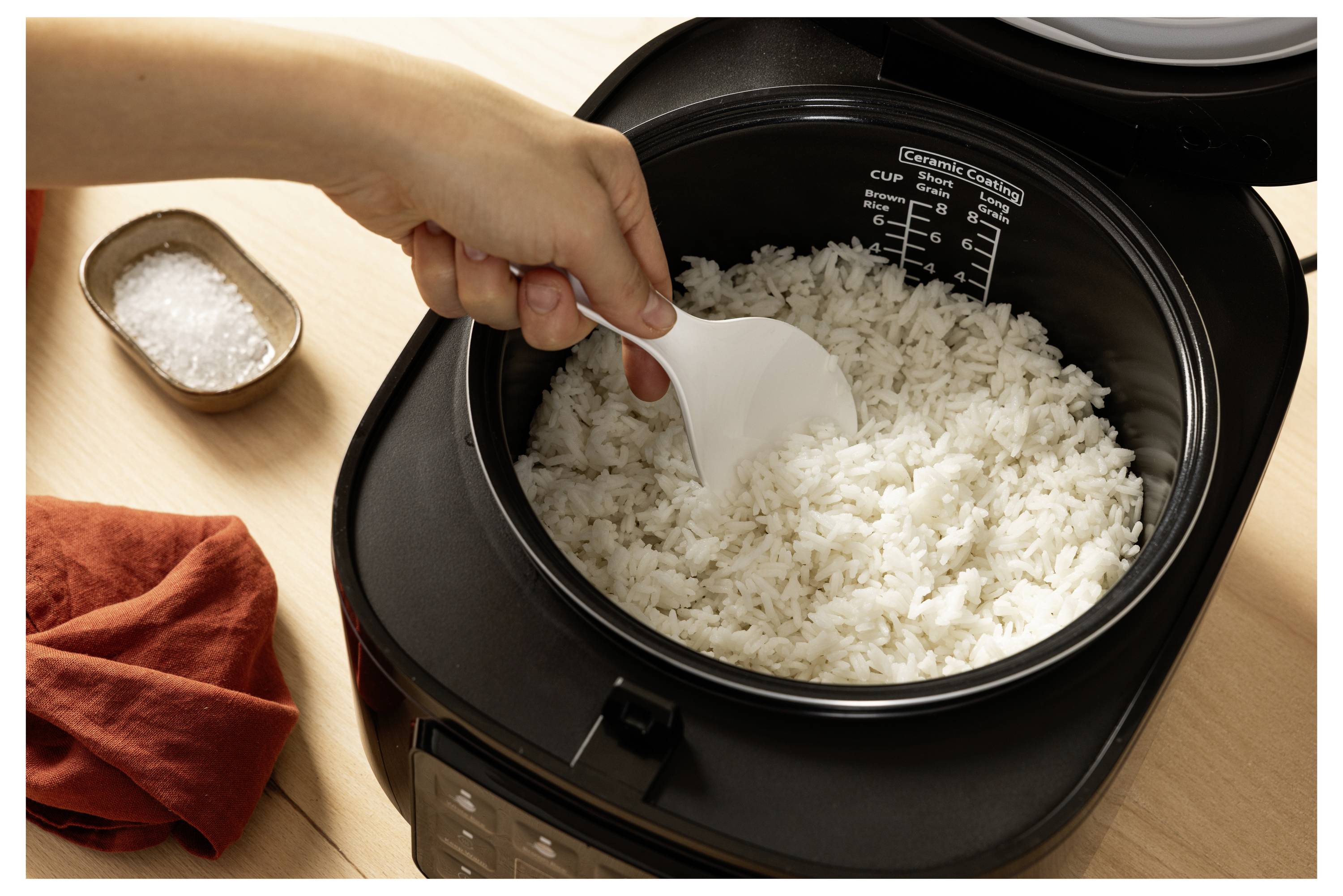 A hand uses a spatula to scoop cooked white rice from an electric rice cooker, with a small dish of salt and a red cloth nearby.