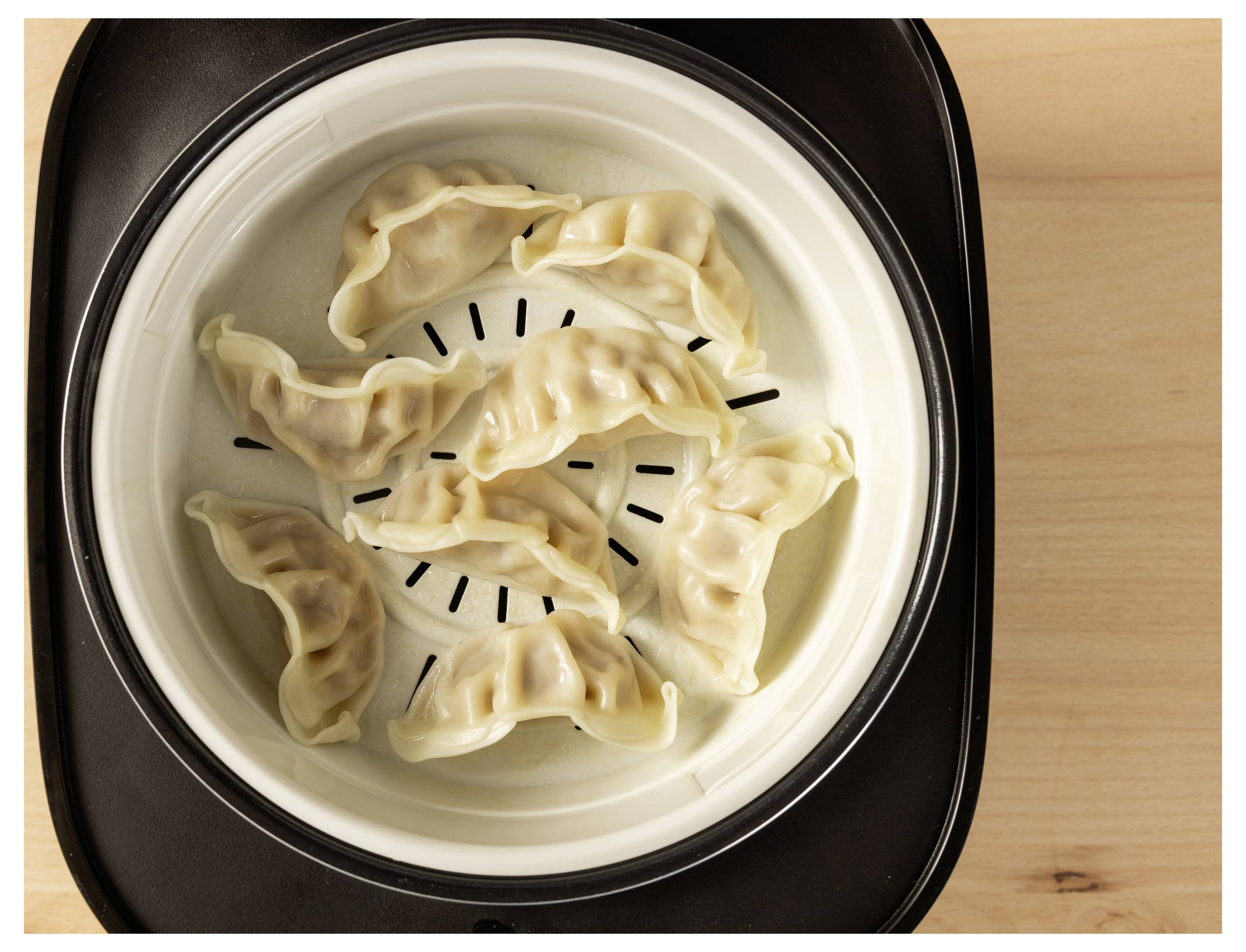Seven dumplings are arranged in a white steamer basket, ready to be cooked. The basket sits on a wooden surface.
