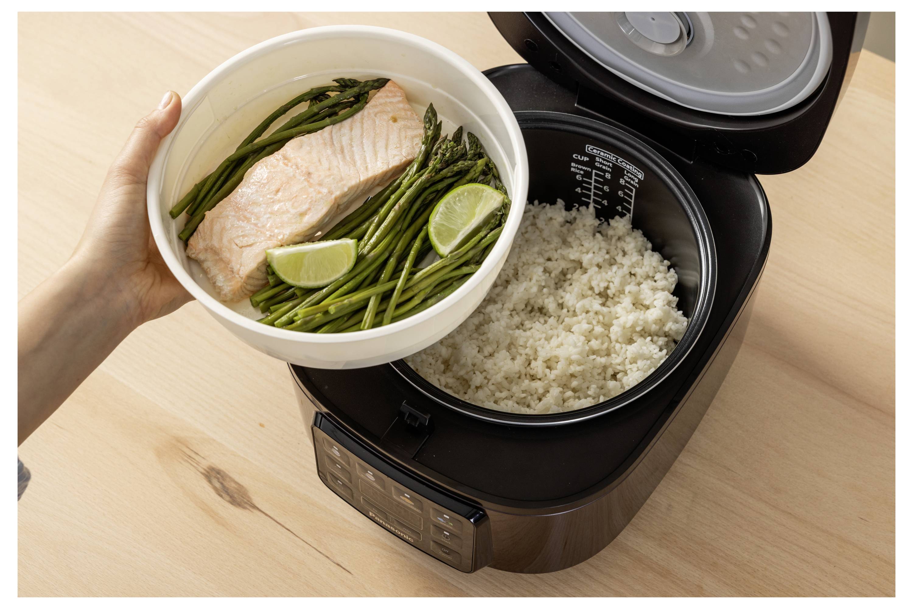 A person holds a plate with cooked salmon, asparagus, and lime slices above a rice cooker filled with rice on a wooden table.