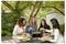Four women enjoy cooking and eating at an outdoor table surrounded by greenery. A grill is placed on the table.