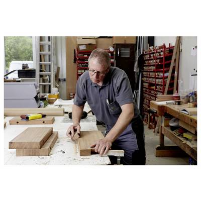 A person working in a carpentry shop, focused on measuring a piece of wood on a large table, surrounded by tools and equipment.
