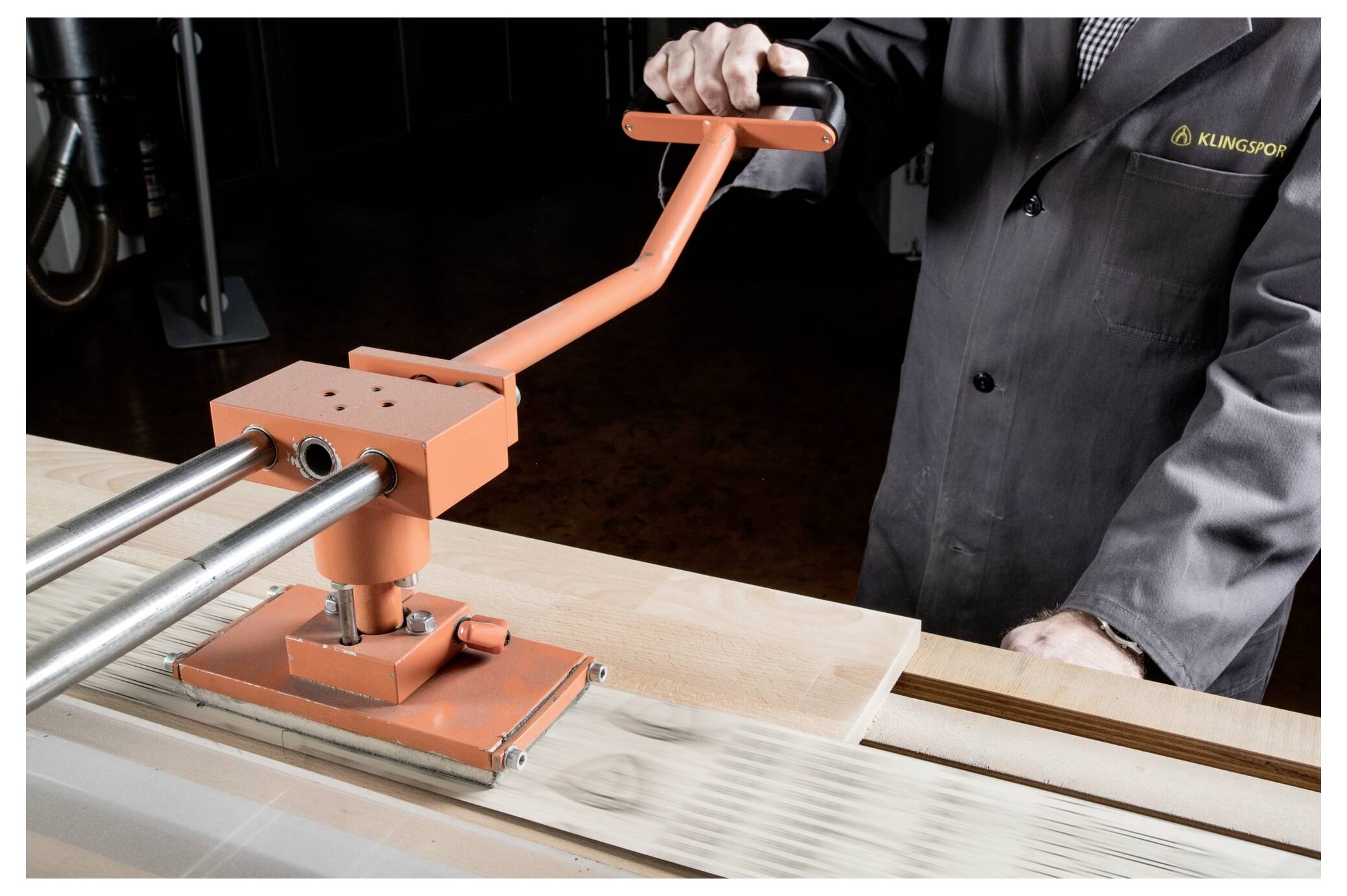 A person operates a manual tile cutter, aligning tiles for cutting on a workbench. The cutter has a long handle and two metal guide rails.