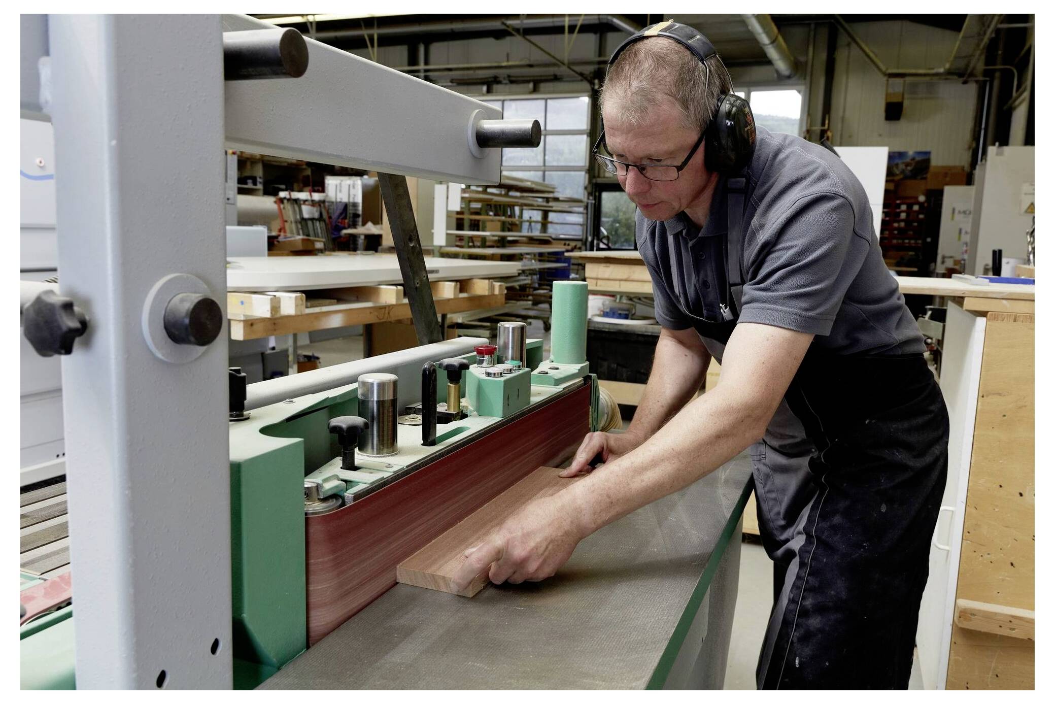 A worker smooths a wooden board on a large sanding machine in a workshop, wearing safety glasses and earmuffs for protection.