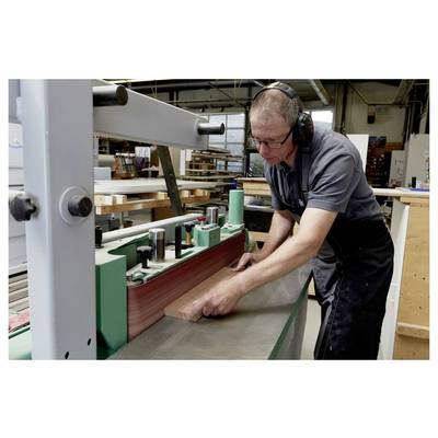A person wearing safety gear operates a sanding machine in a workshop, smoothing a piece of wood with focused attention.