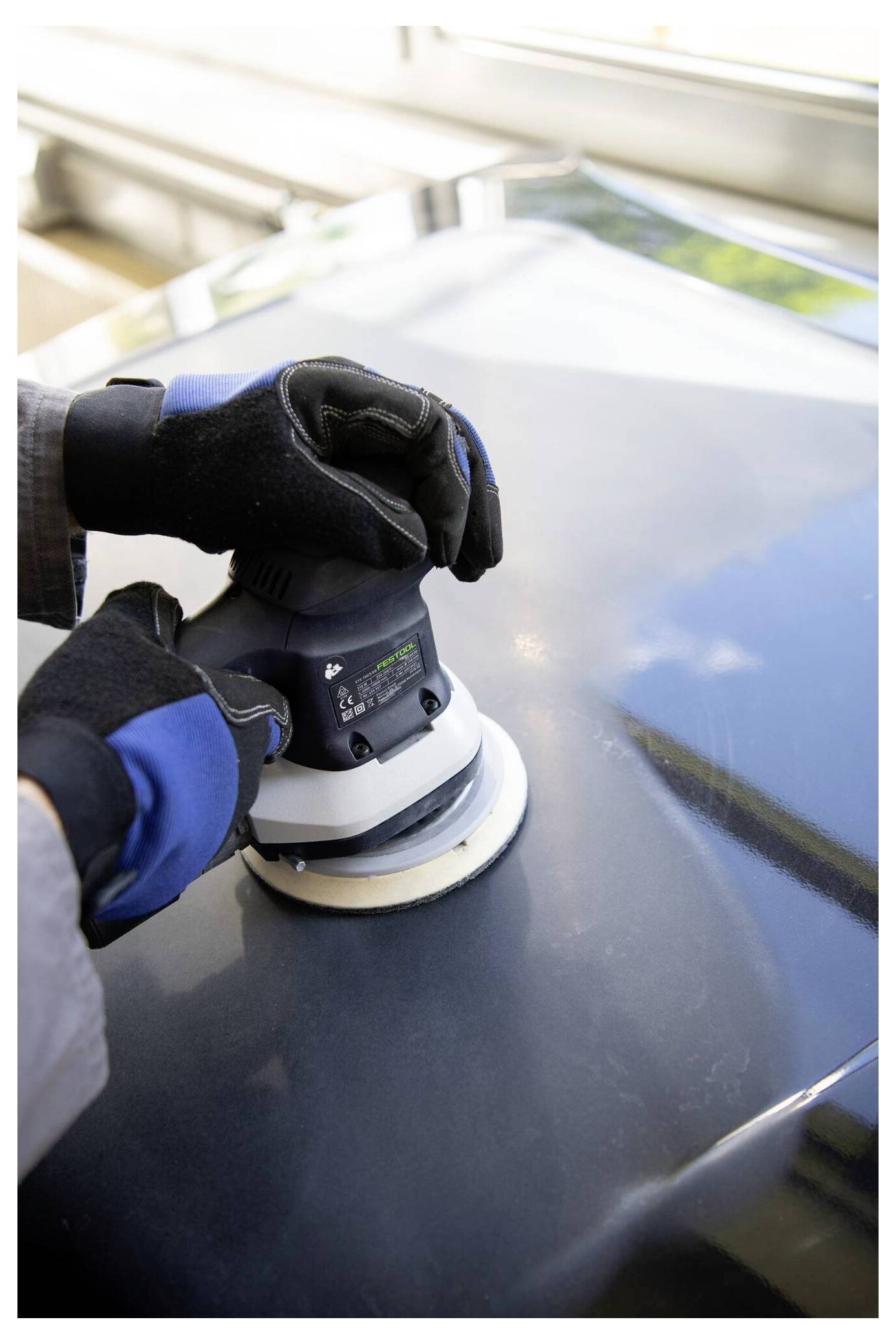 A person wearing gloves uses an electric polisher on a glossy, dark surface, likely a car hood, under natural light.