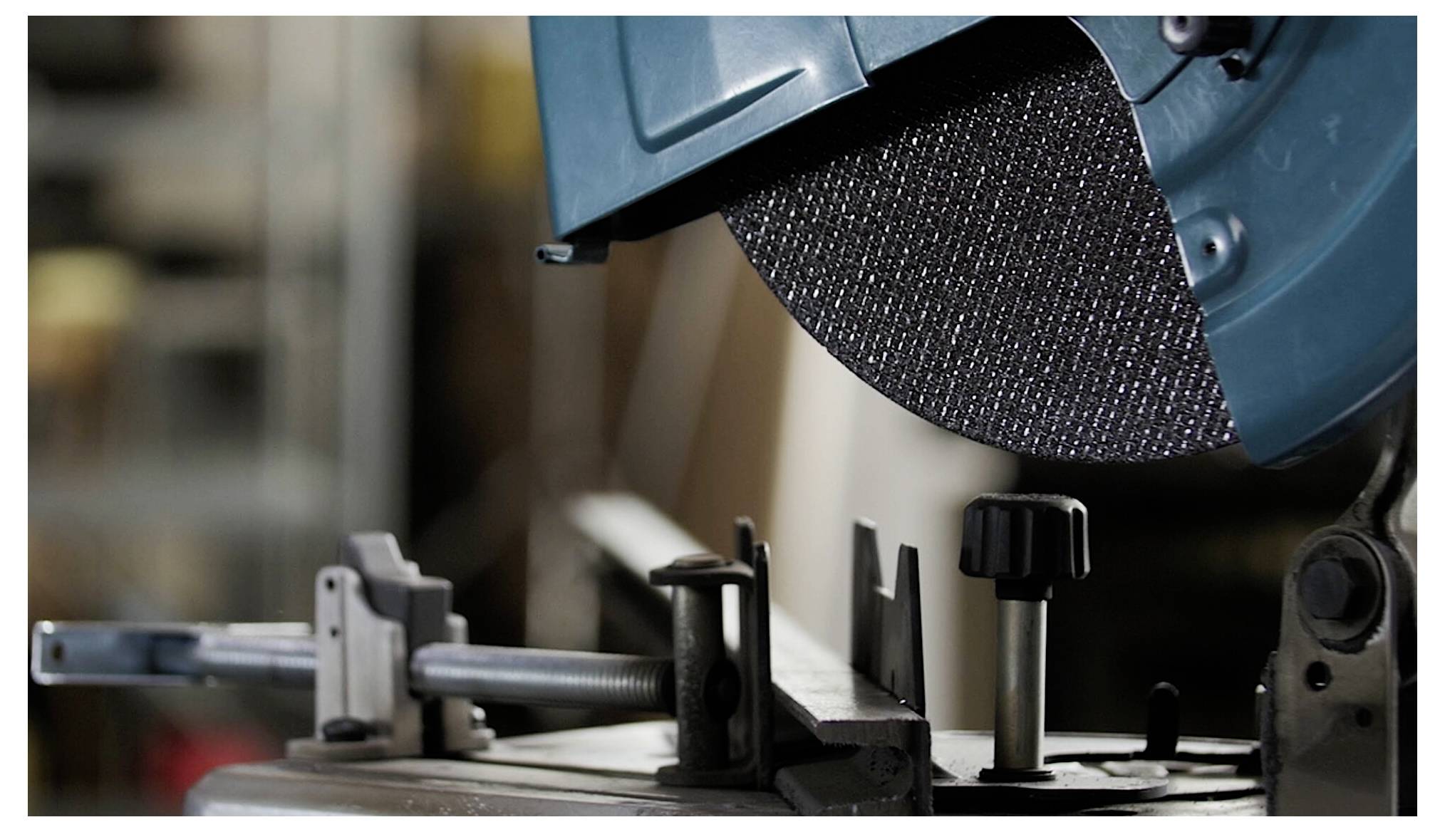 A close-up of a circular saw blade in a workshop, partially enclosed in a protective cover, with a metal clamping device in the foreground.