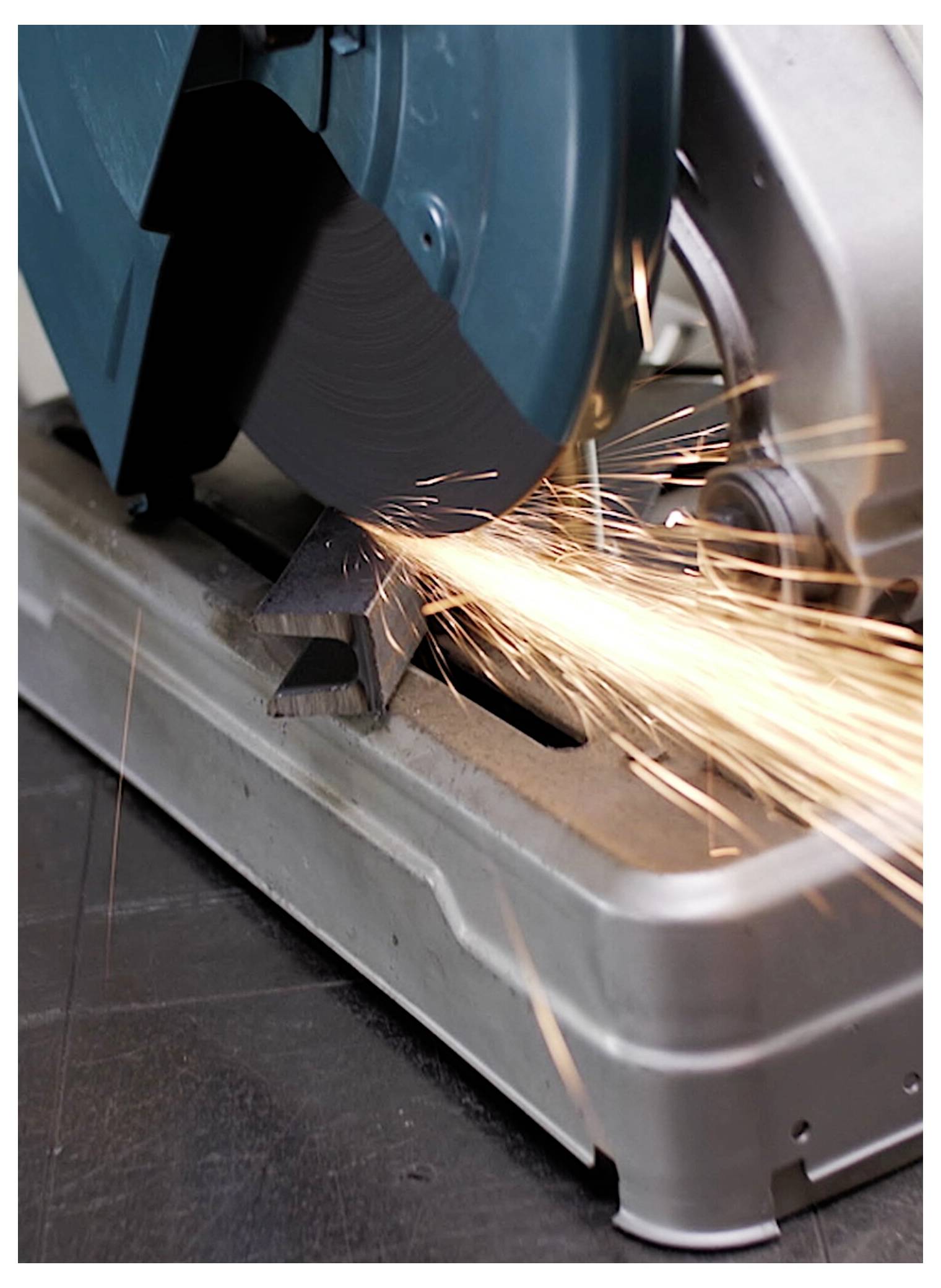 A metal cutting saw with blue circular blade in motion, producing bright orange sparks as it cuts through a metal bar on a workshop floor.
