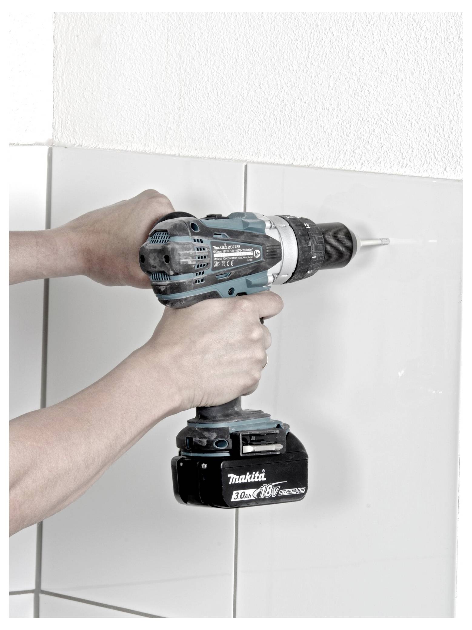 A person uses a cordless drill to make a hole in a white tiled wall.