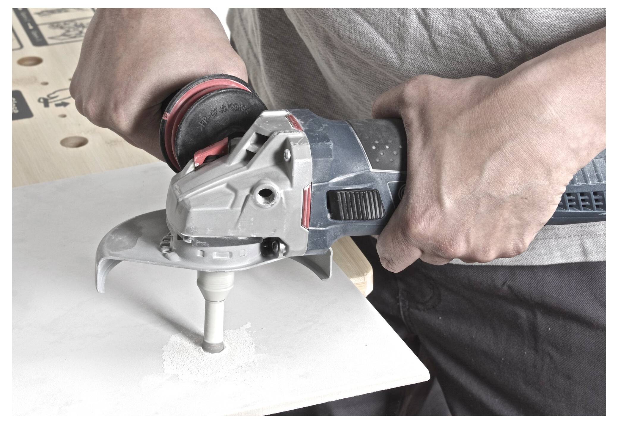 A person uses an electric tool to drill a hole in a white tile. The tool has a circular attachment and is held with both hands.